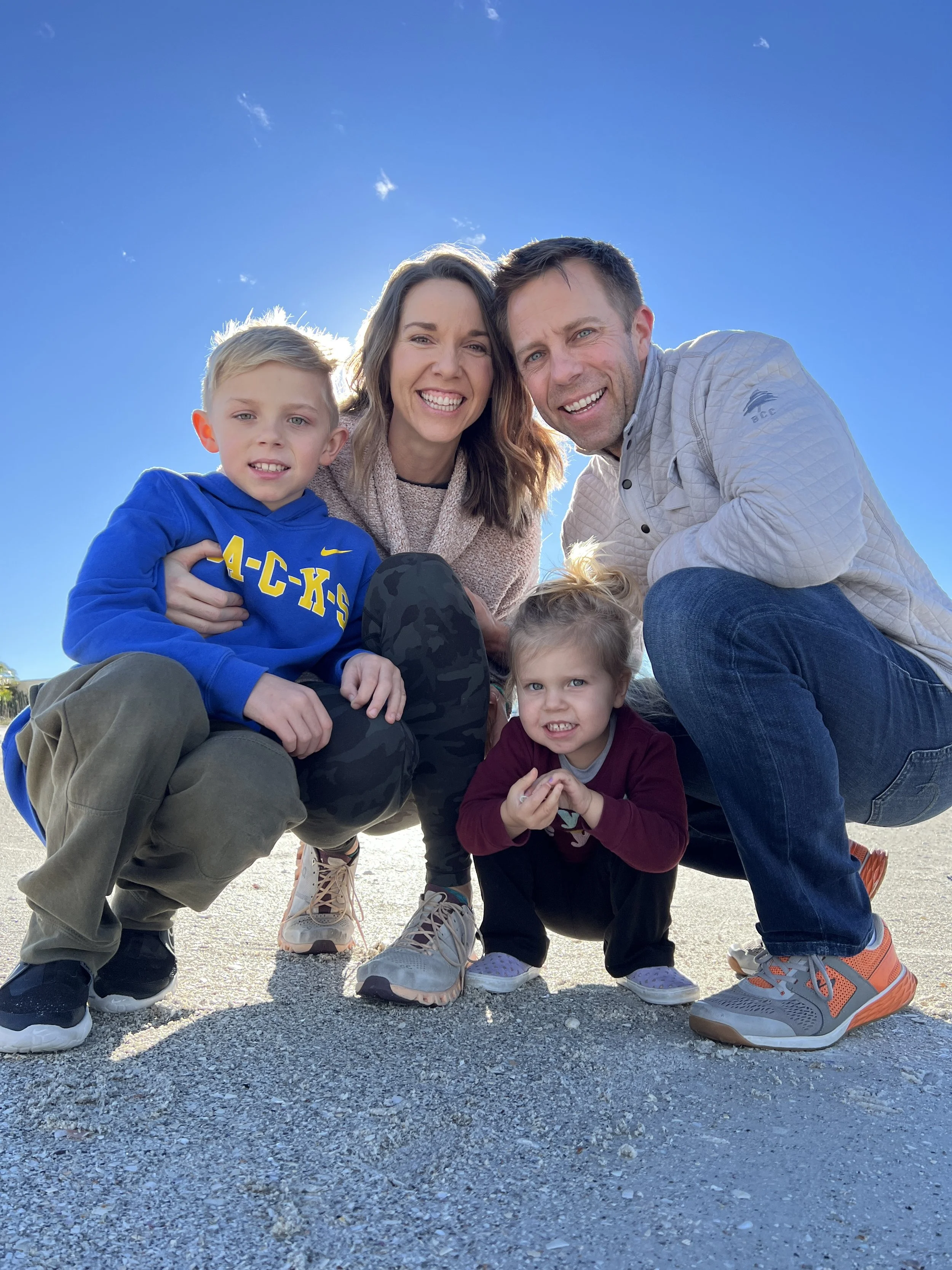 Family of four posing outdoors on a sunny day, smiling at the camera. The family includes a woman, a man, a young boy, and a young girl, all kneeling or squatting on the ground with a clear blue sky in the background.