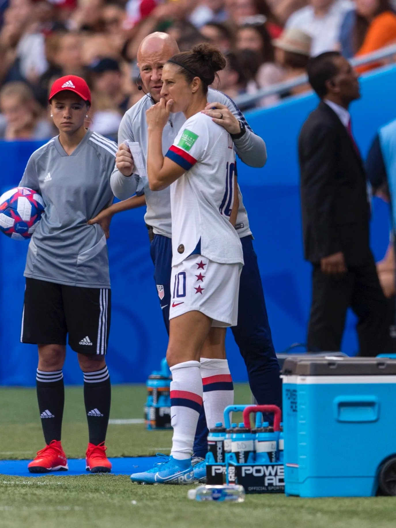 A female soccer player dressed in a white US national team uniform with the number 10, standing on the sideline with her hand on her chin, listening to a coach. There is a water cooler, water bottles, and medical supplies nearby.