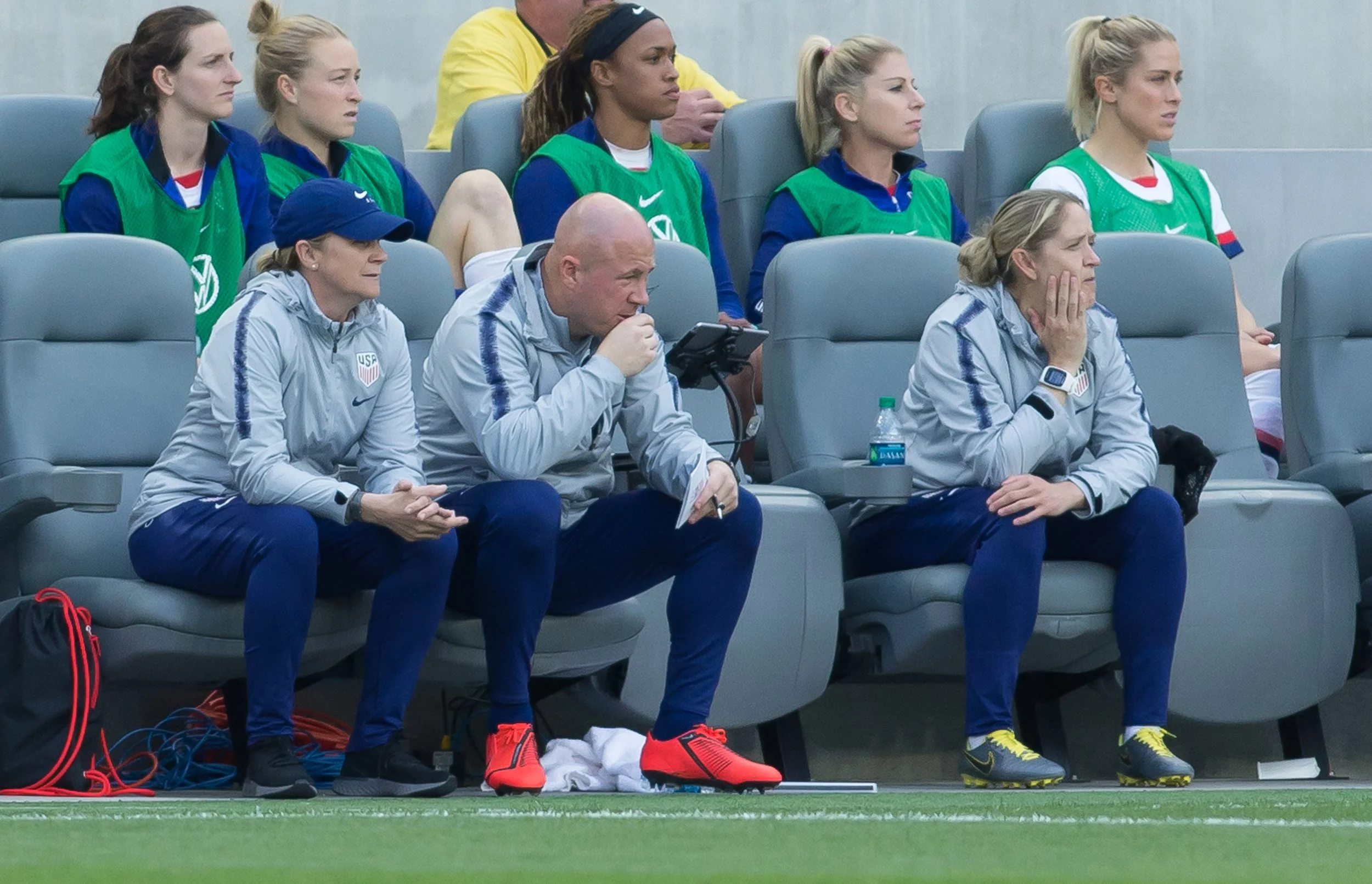 Female soccer players and coaching staff sitting on the bench, focused on a match. The players wear green and white bibs, while the staff are in gray jackets, with some holding phones or watching the game intently.