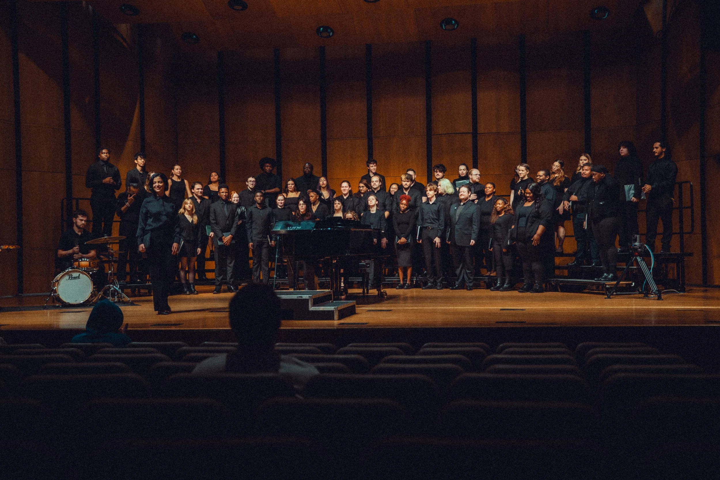 A choir of diverse people dressed in black on stage, standing in front of a wooden wall. There is a pianist, a drum set, and a conductor among them. Audience members are visible in the foreground.