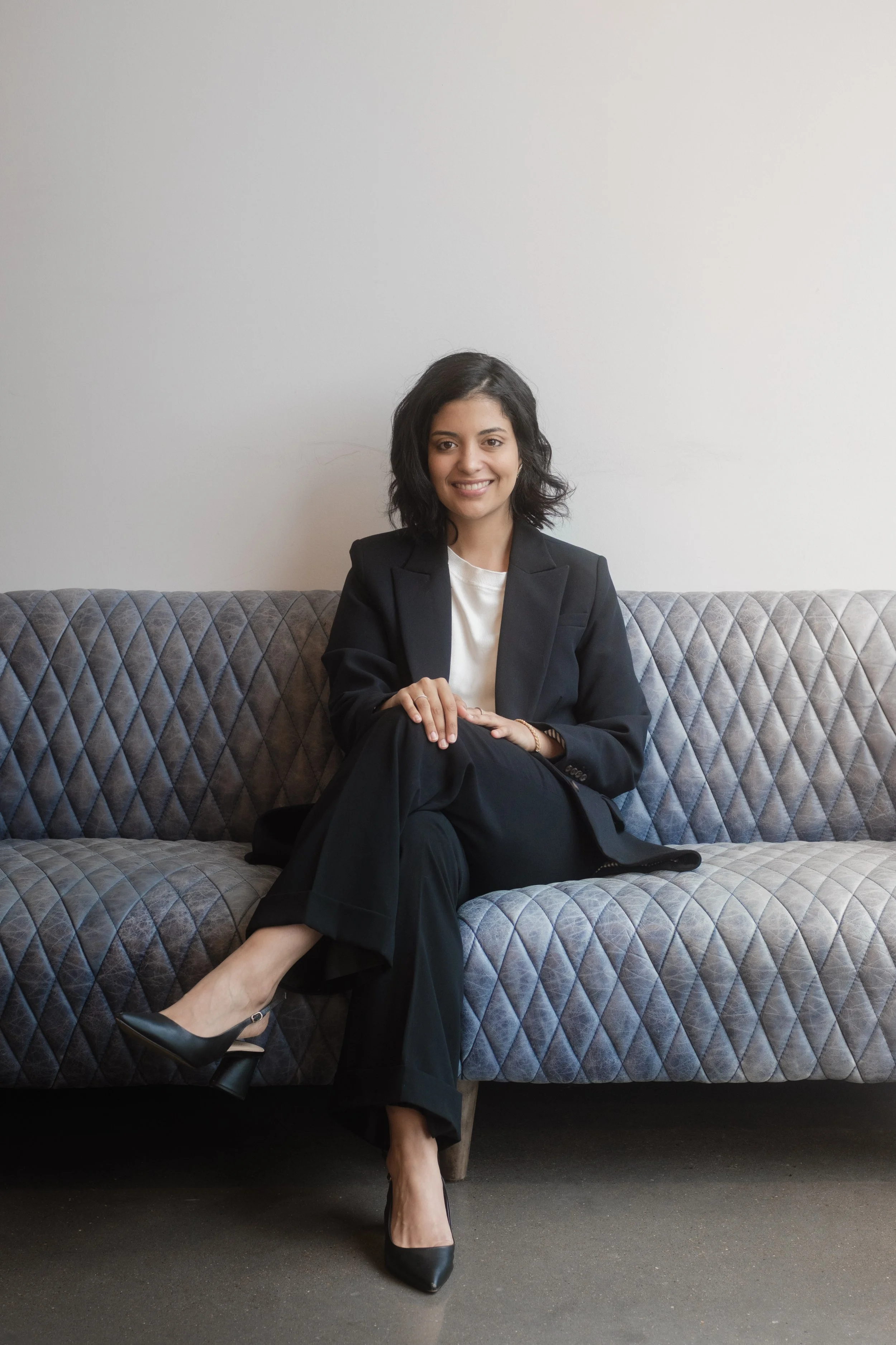 A woman in a black suit sitting on a quilted gray sofa, smiling, with legs crossed and black heels, against a plain white wall.