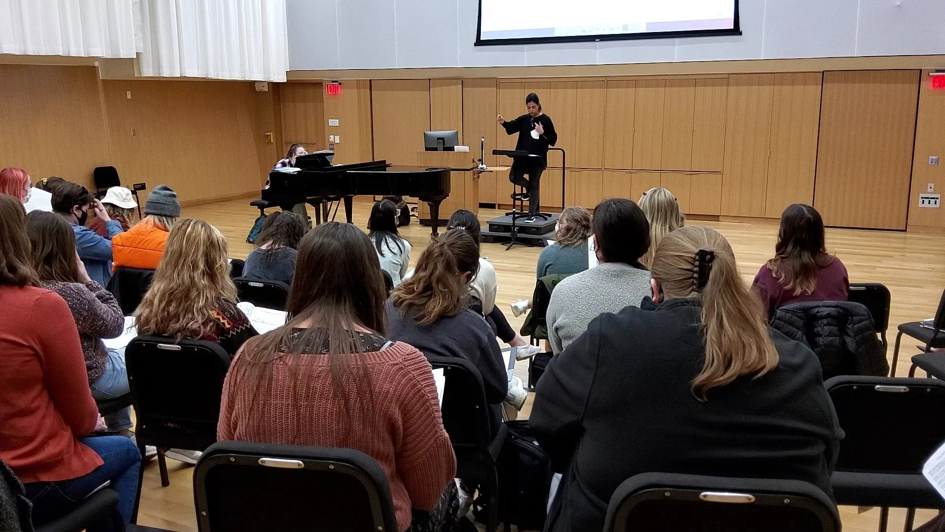 A woman conducting a music class with a piano and a music stand, in front of an audience of people coaching note sheets.