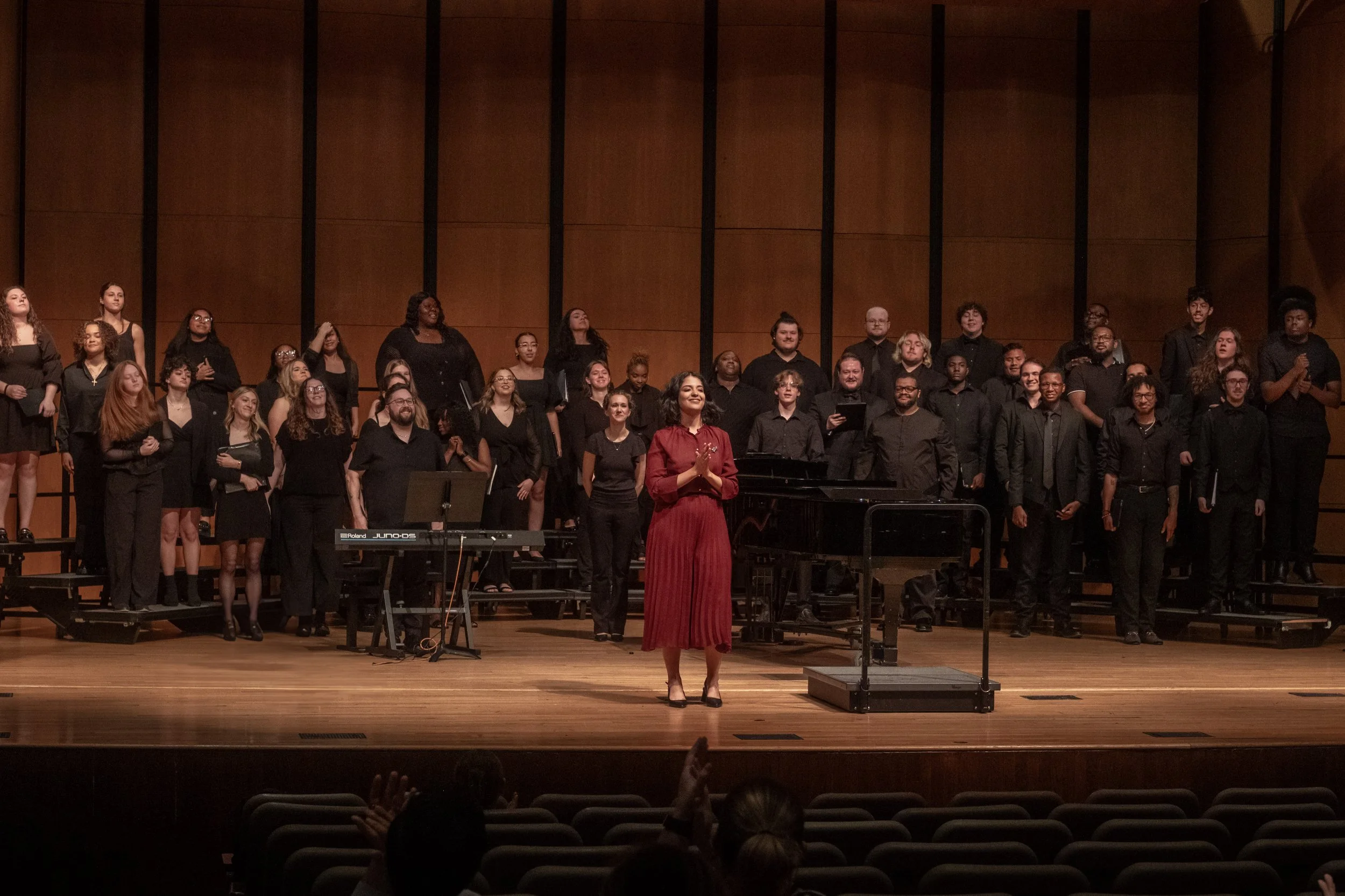 A group of singers on stage with a woman in a red dress at the front, clapping, in a concert hall with wooden paneling.