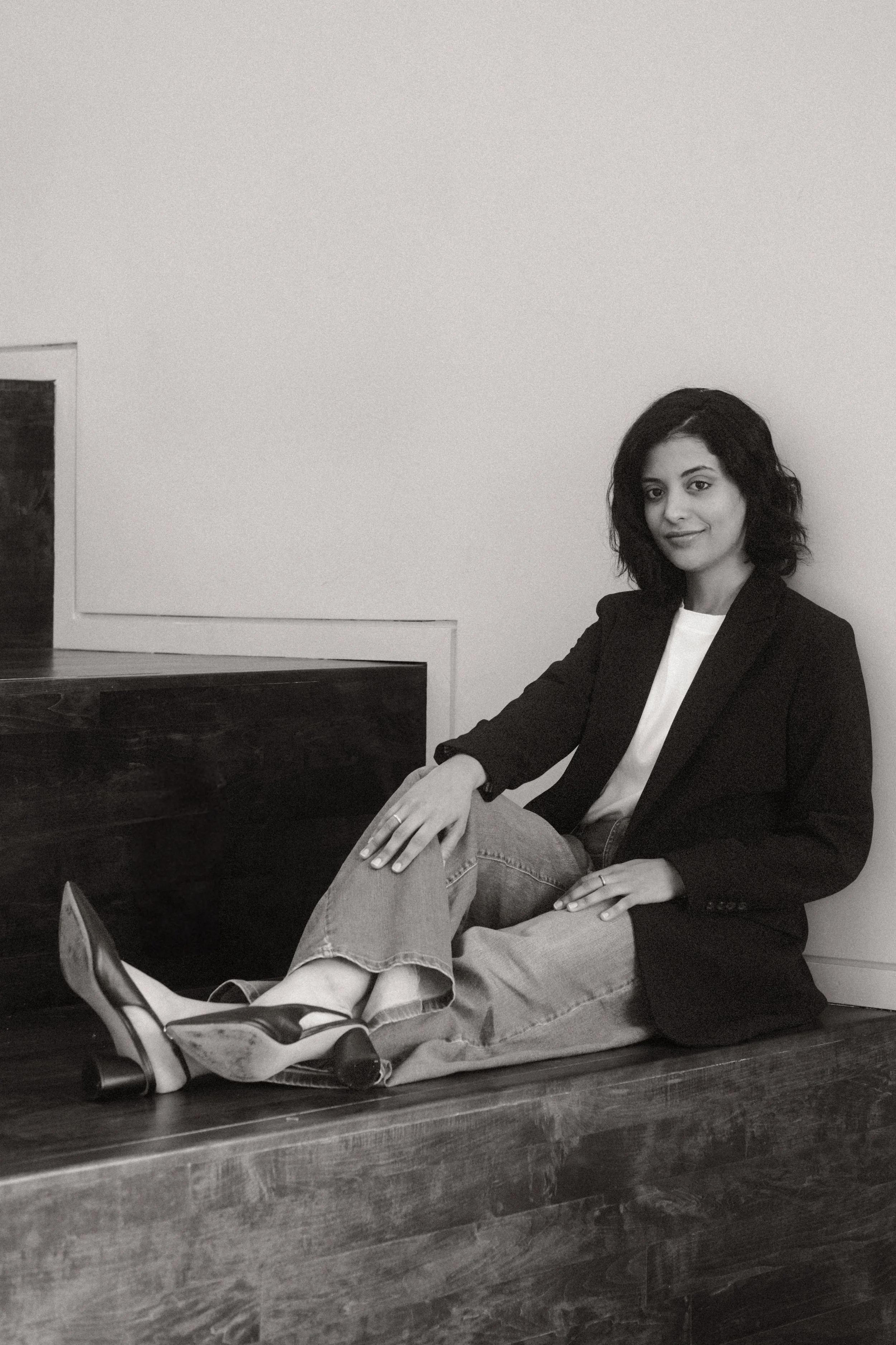 A woman with dark, curly hair sitting on a wooden surface, wearing a blazer, jeans, and heels, with a relaxed expression, against a plain wall.