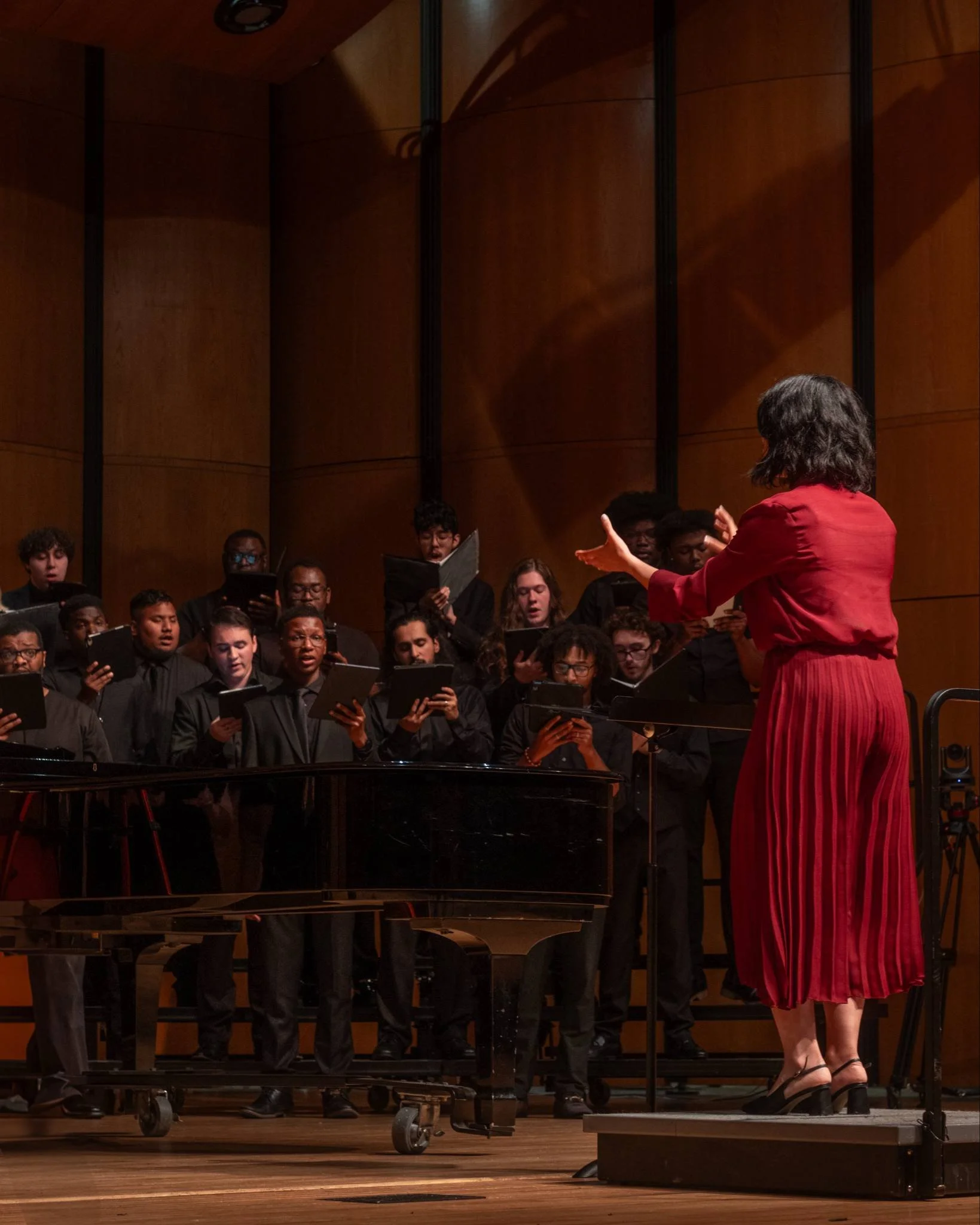 A choir performing on stage, directed by a woman in red who is conducting with her hands, with a grand piano in front of the choir and a wooden backdrop.