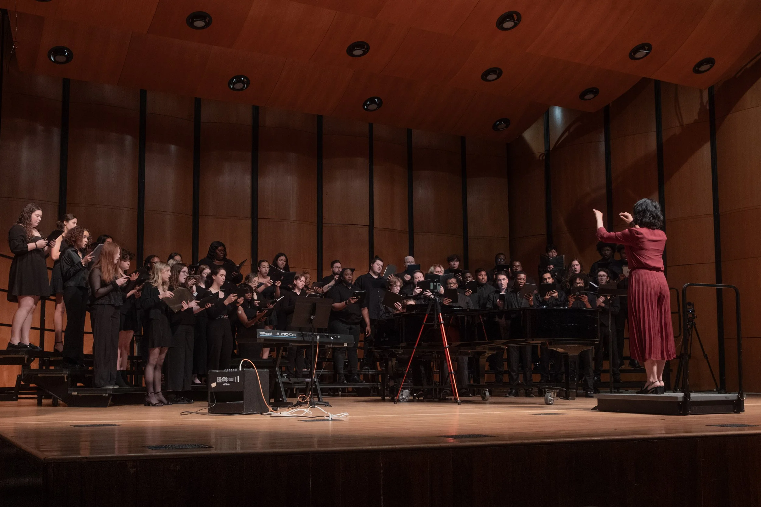 A choir of people wearing black standing on risers on a wooden stage, led by a conductor in a red dress who is conducting with her right hand, inside a wood-paneled concert hall.