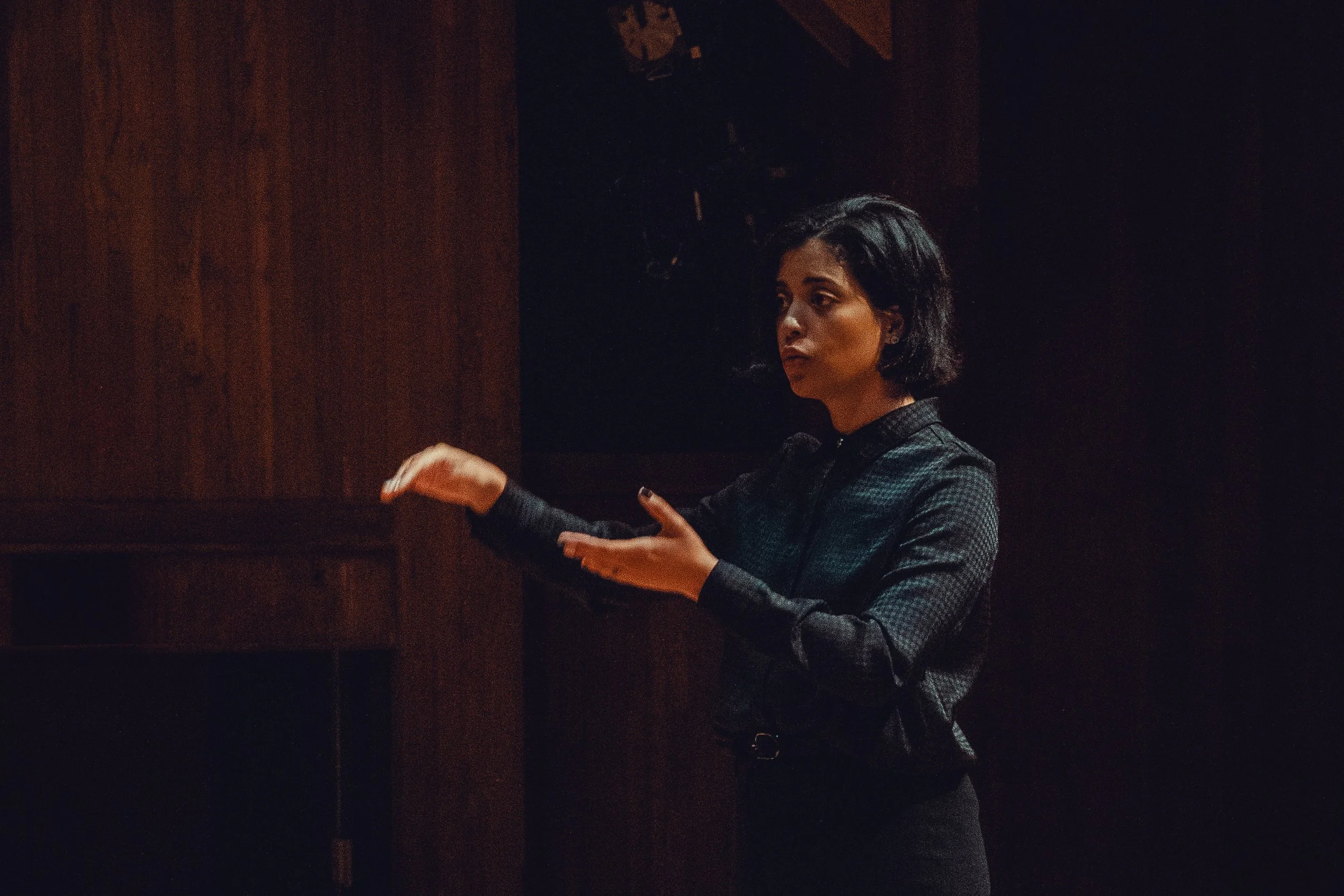 A woman with short black hair, wearing a dark, long-sleeved blouse, stands against a dark wooden background, gesturing with her hands as if speaking or explaining something.