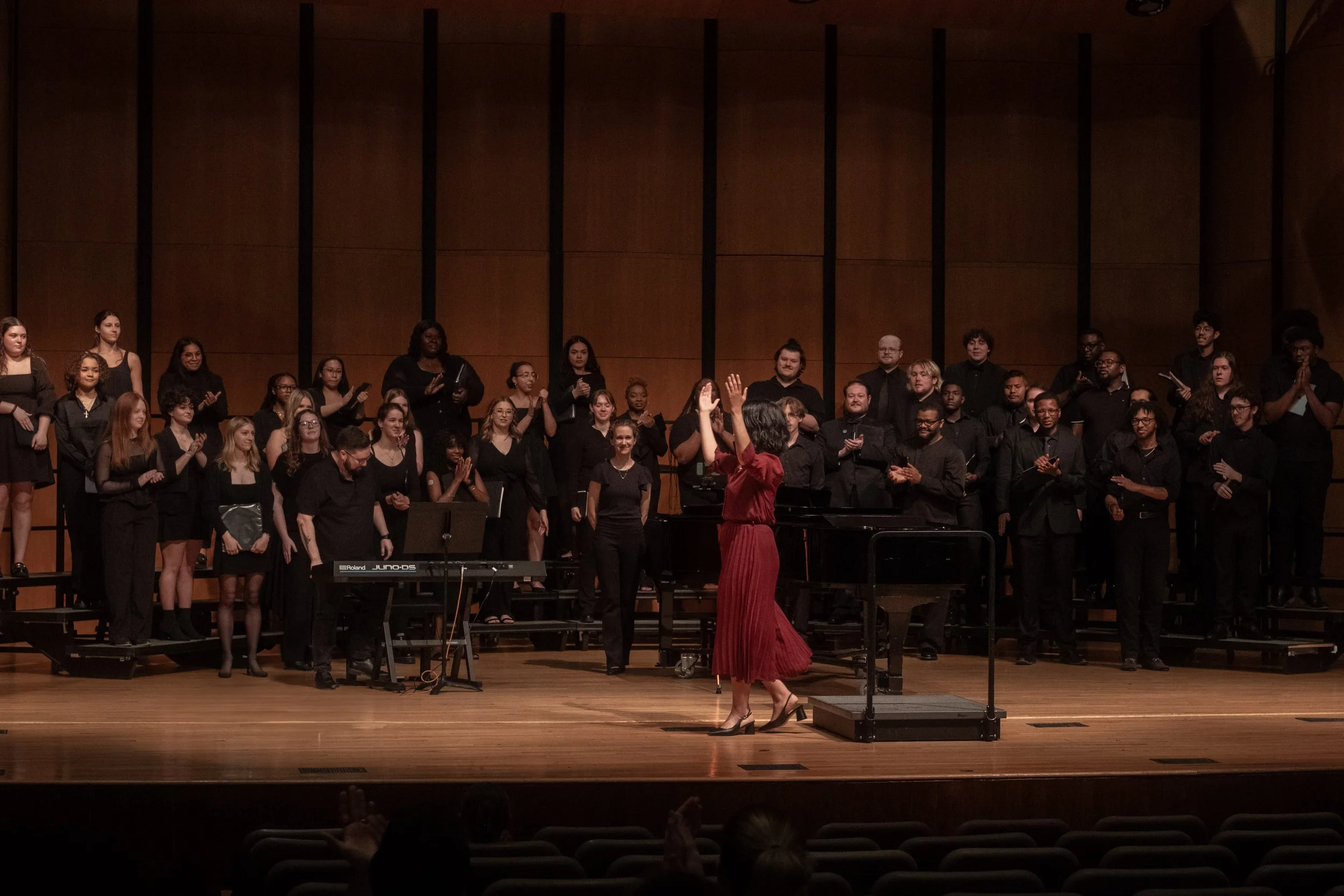 A choir standing on stage with a woman conductor in a red dress leading them, during a performance in a concert hall with wooden paneling.