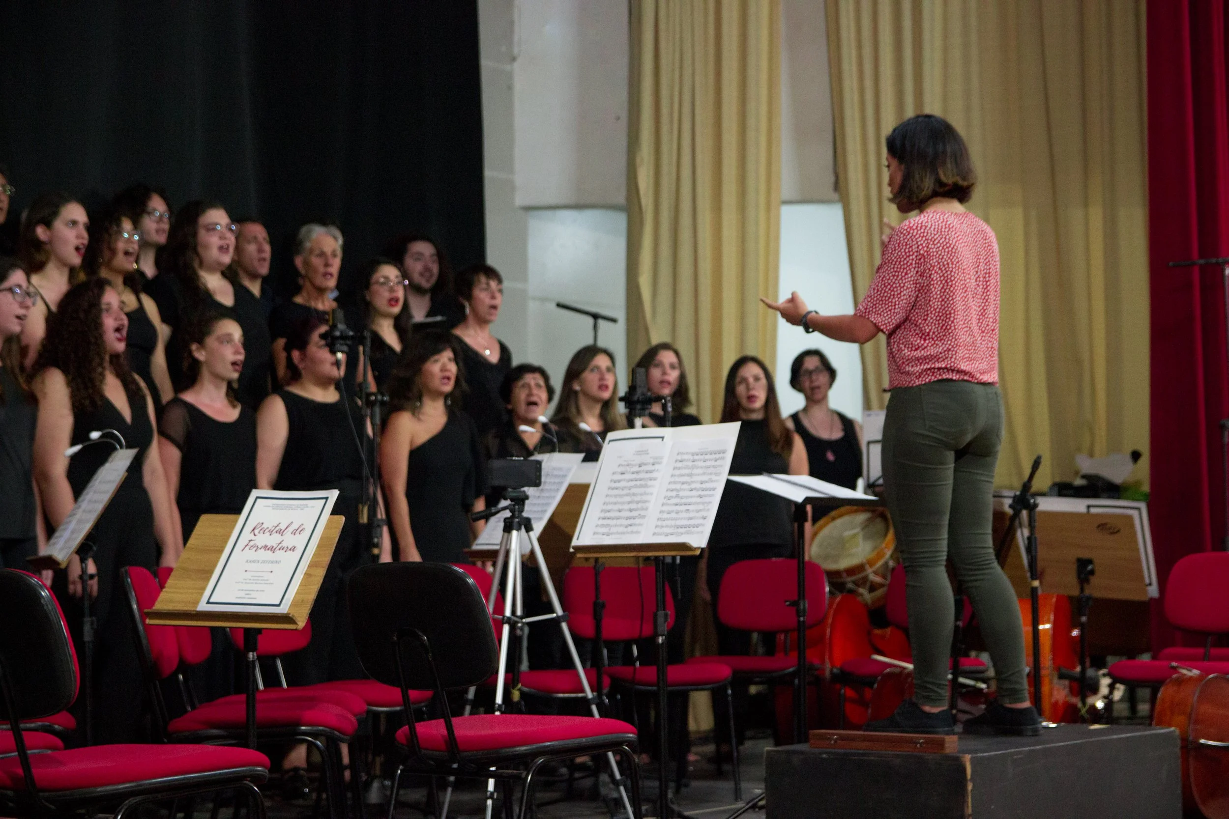 Choir performing during rehearsal or concert, directed by a conductor standing on a raised platform, with sheet music stands and colorful curtains in the background.