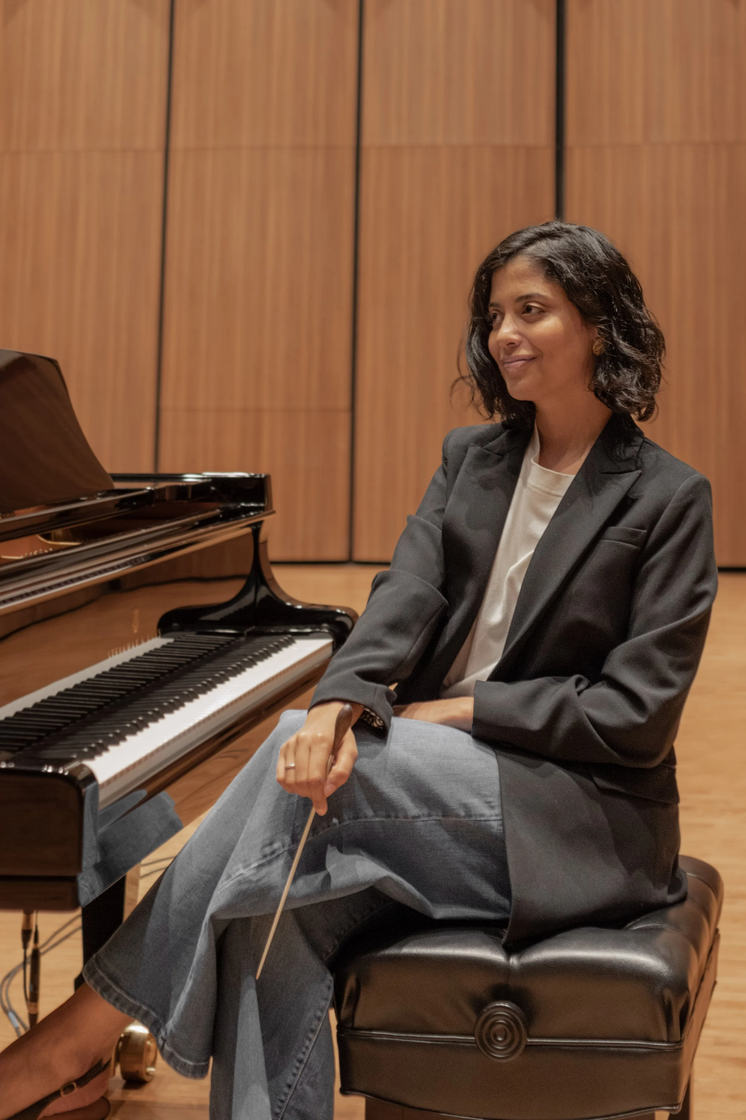 A woman in a black blazer and gray pants sitting on a piano bench in front of a grand piano in a music studio or concert hall.