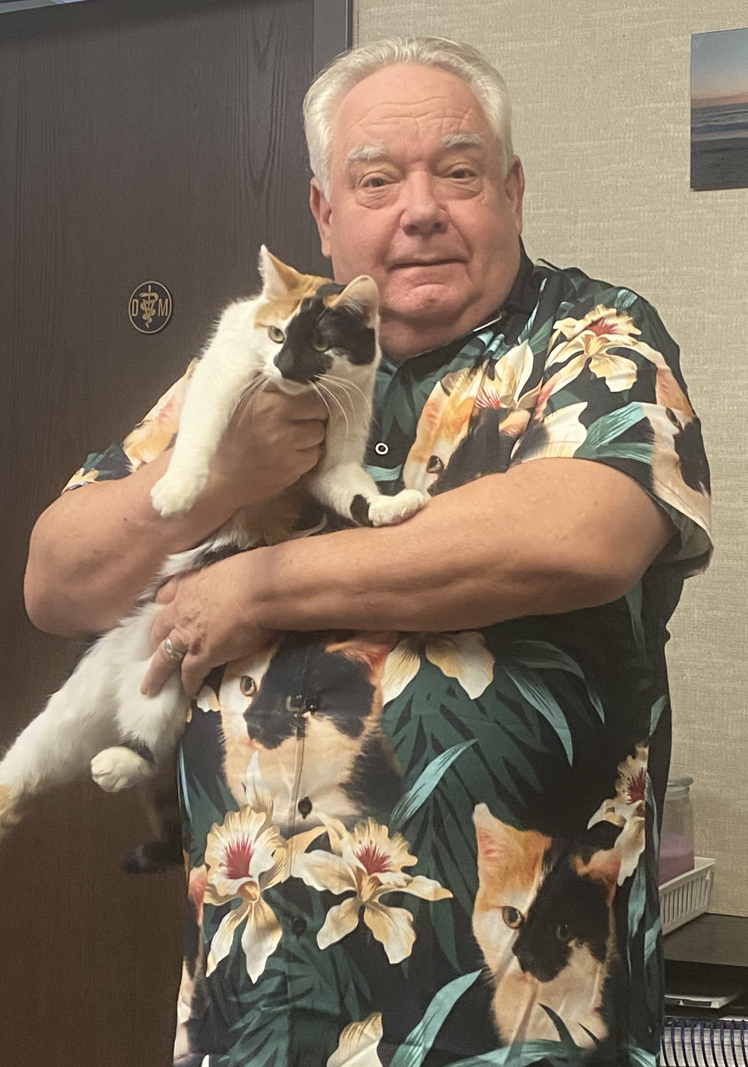 An elderly man with white hair wearing a floral Hawaiian shirt, holding a calico cat with one paw hanging down and a relaxed expression in an indoor setting.