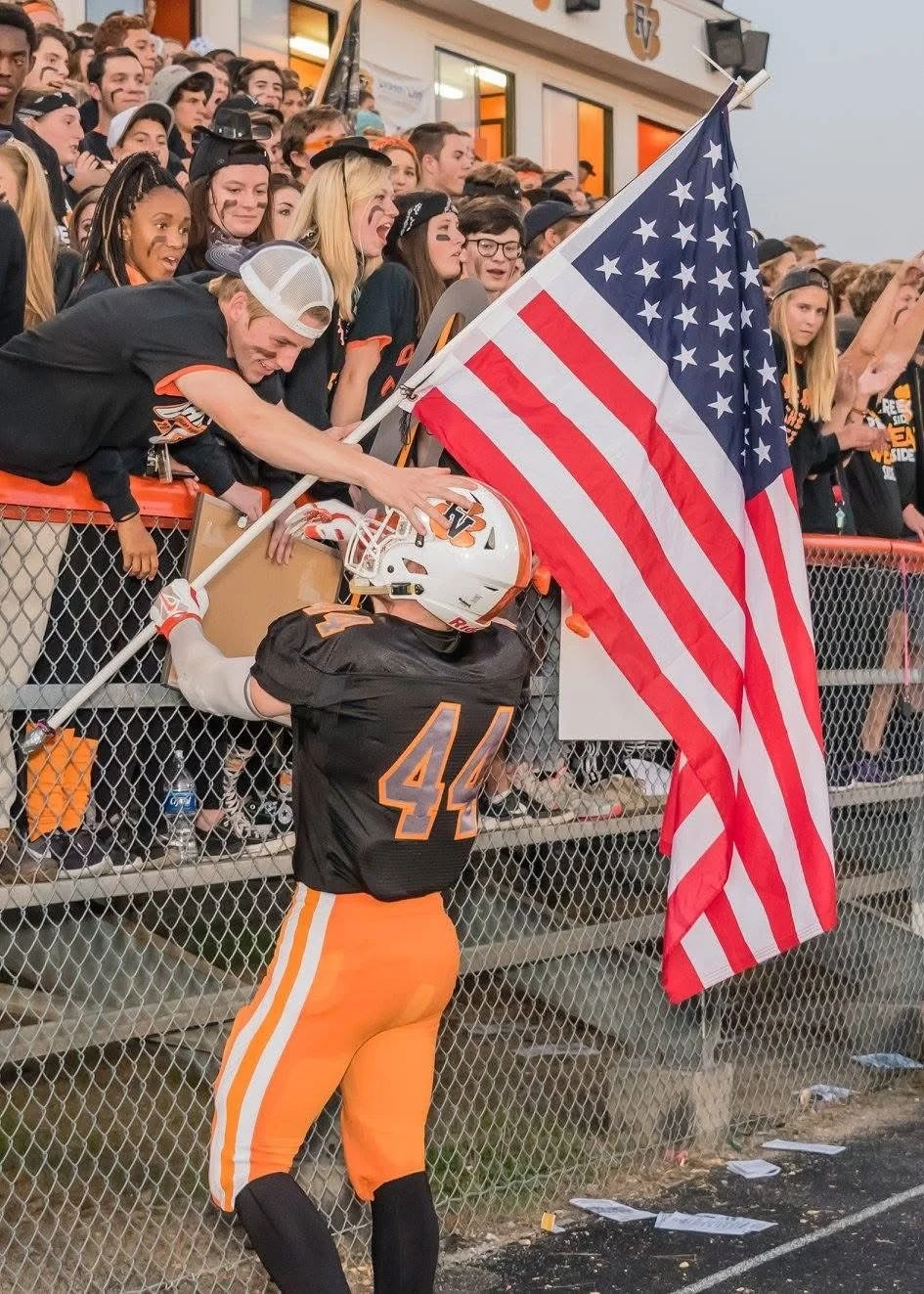 A football player in a black and orange uniform exchanges a flag with a fan over a fence, with a crowd of fans in the background.