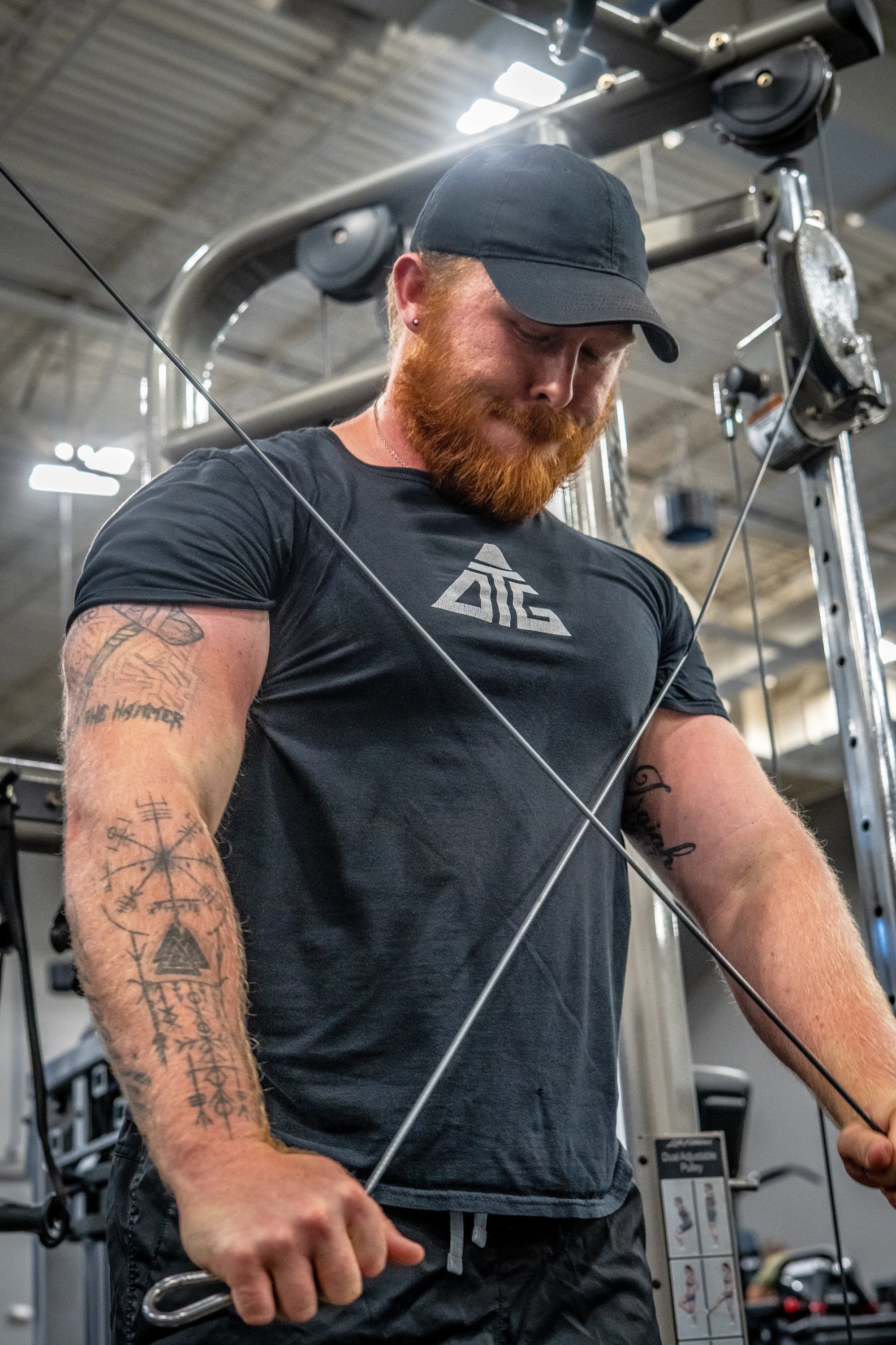 A man with a red beard wearing a black baseball cap and black shirt is performing a cable exercise in a gym.