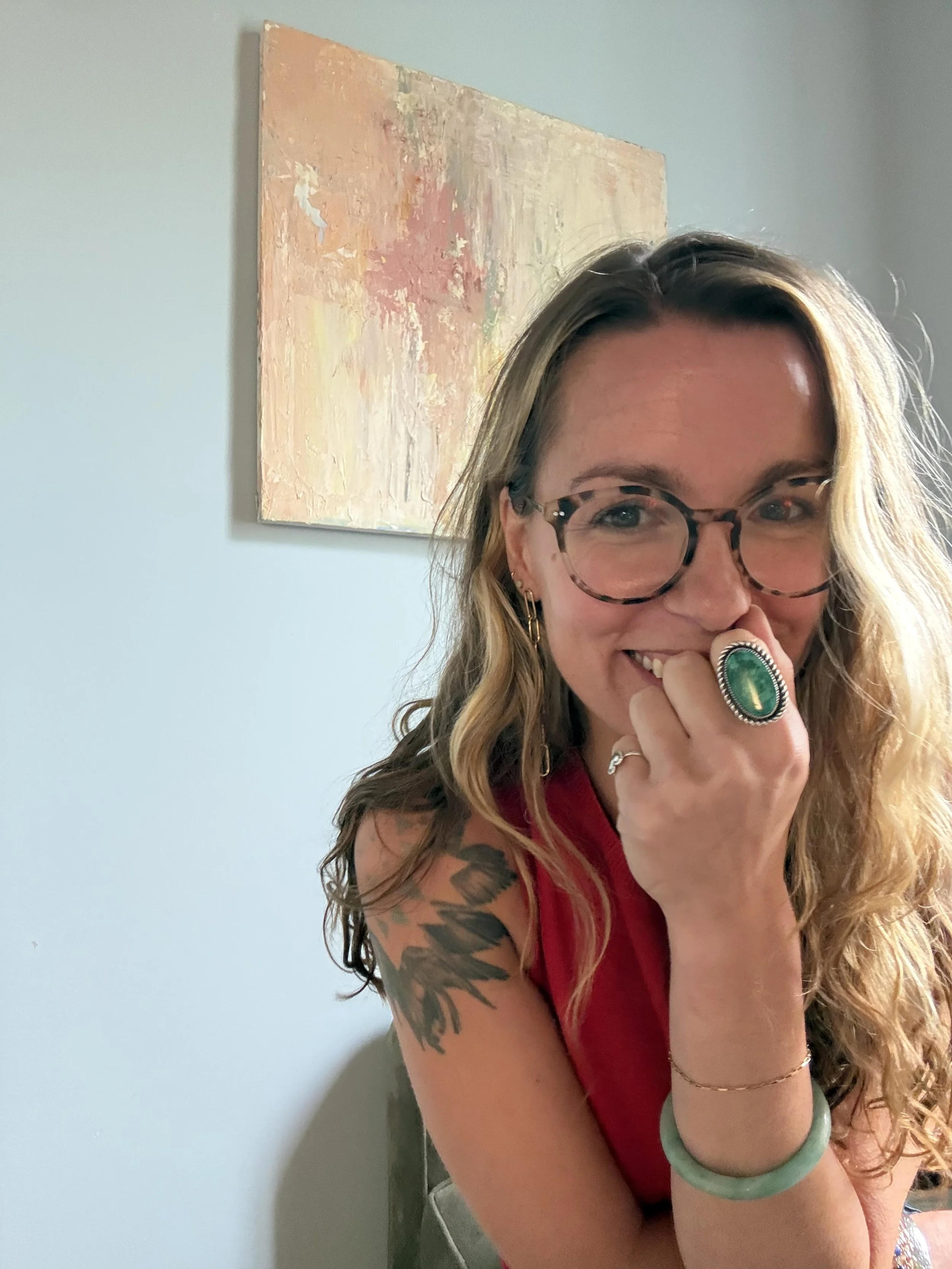 A woman with long wavy hair and glasses, smiling with her hand near her face, wearing a red sleeveless top, and a teal bracelet, with a colorful abstract painting on the wall behind her.