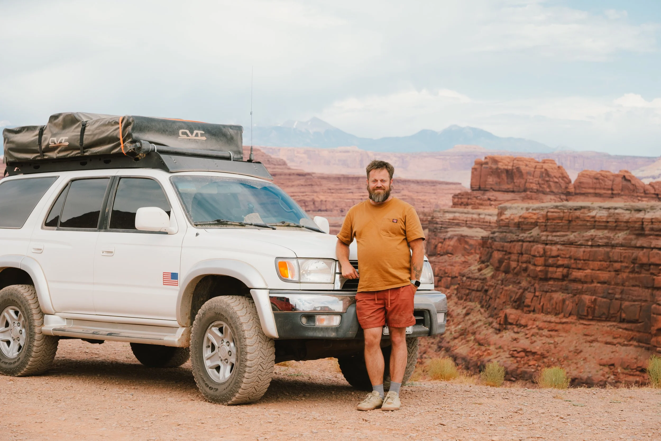 A man with a beard, in an orange t-shirt and shorts, standing next to a white SUV with outdoor gear on the roof, in a landscape of red rock formations and distant mountains.