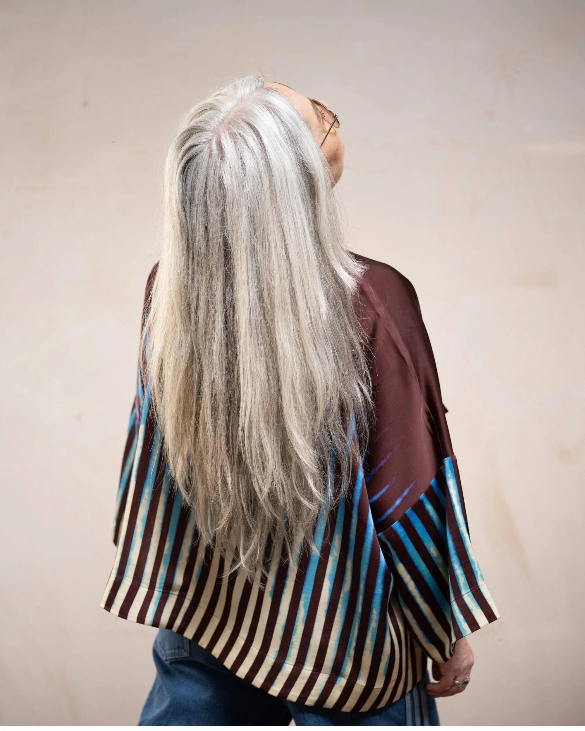 A woman with long, gray hair wearing glasses, a maroon jacket, and striped shirt standing against a plain background.