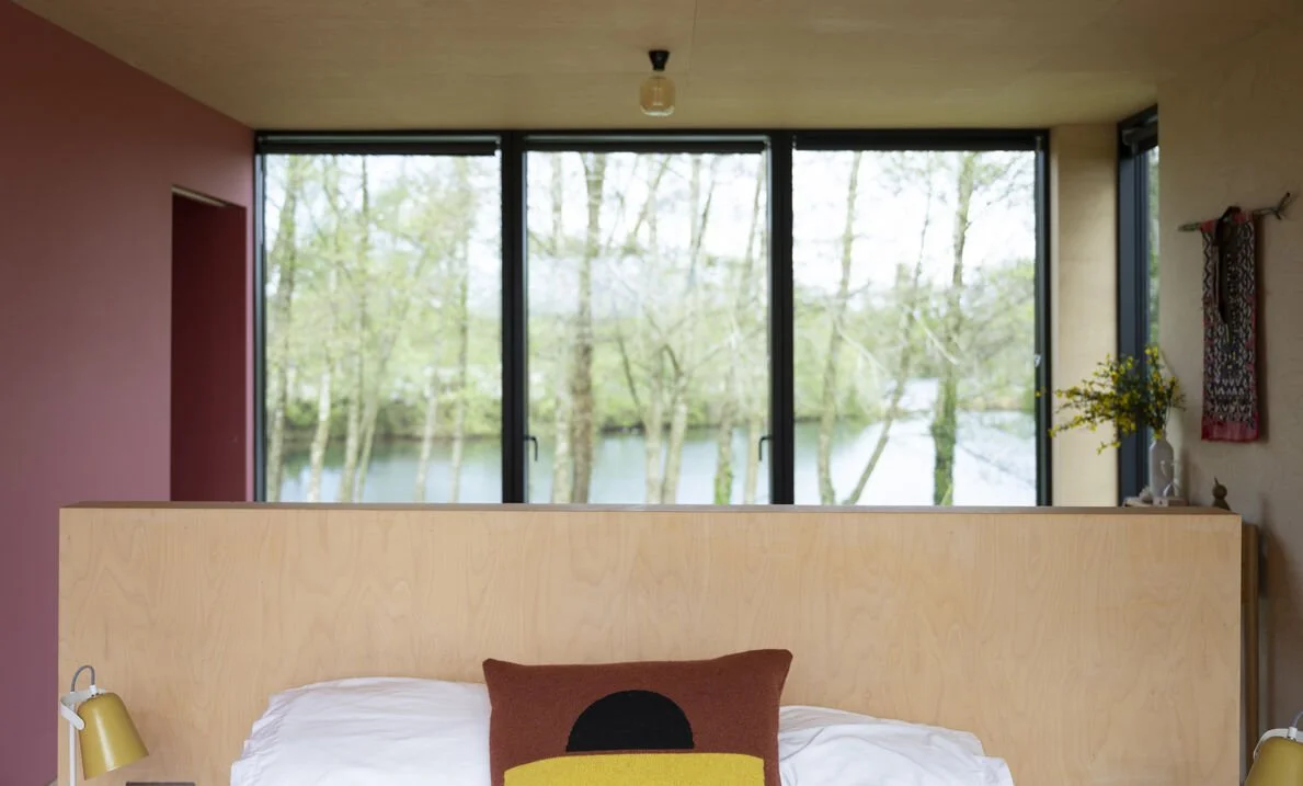 The image depicts a bedroom with a large window overlooking a river and trees outside. The bed is in the foreground with a wooden headboard, a pillow, and a small bedside lamp on the left.