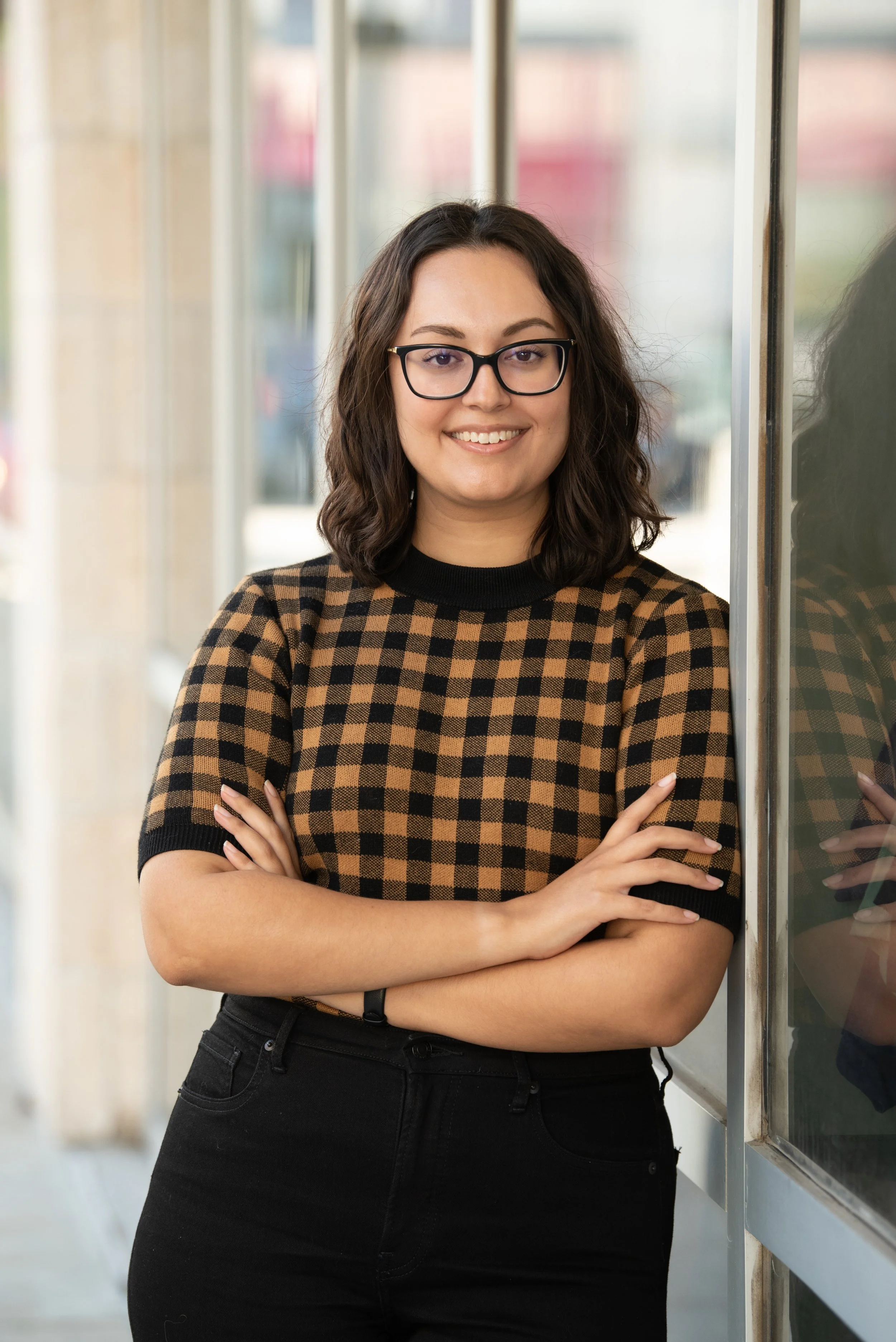 A woman with dark wavy hair, wearing glasses and a black and brown checkered shirt, standing with her arms crossed next to a glass wall.