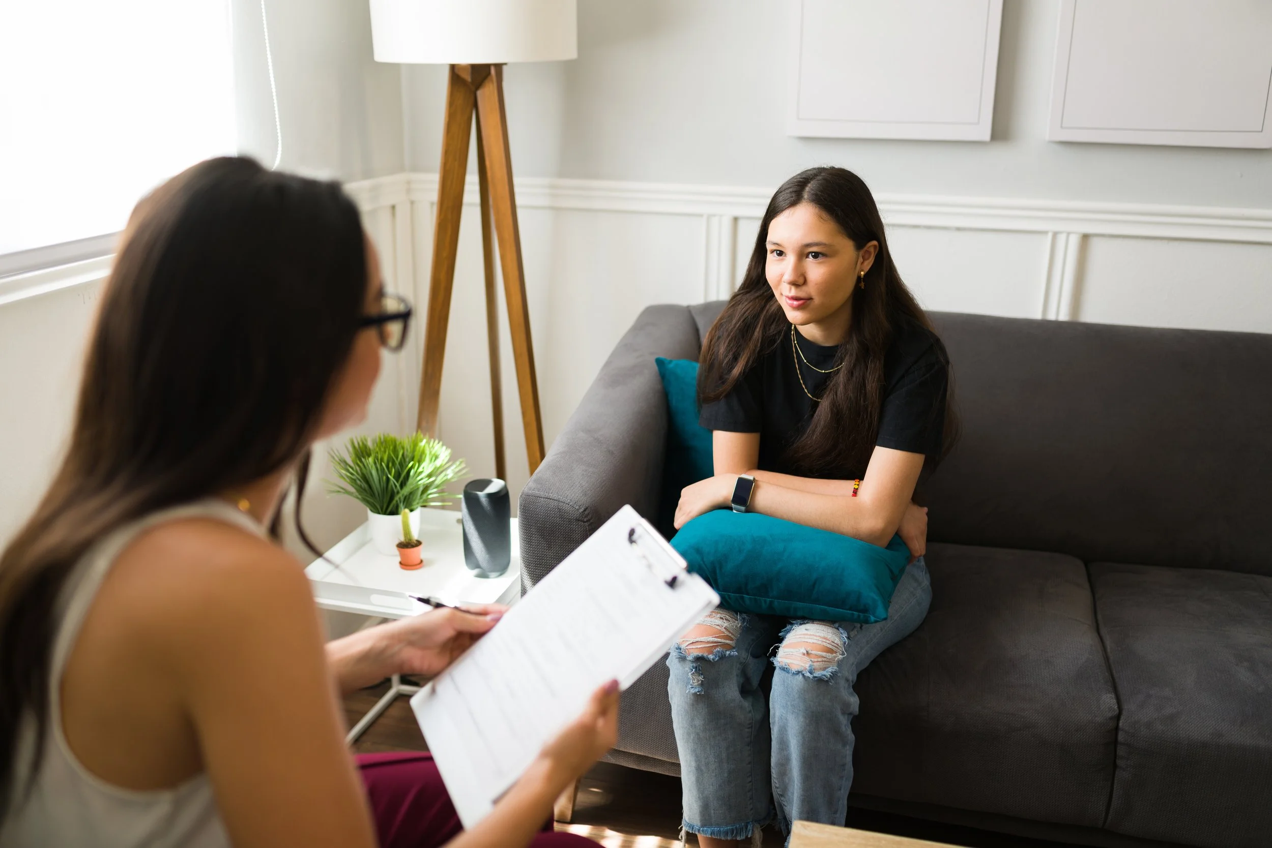A young woman sitting on a couch during a therapy session, talking to a therapist who is holding a clipboard. The room has white walls, a standing lamp, a small white table with a green plant, and a blue cushion on the couch.