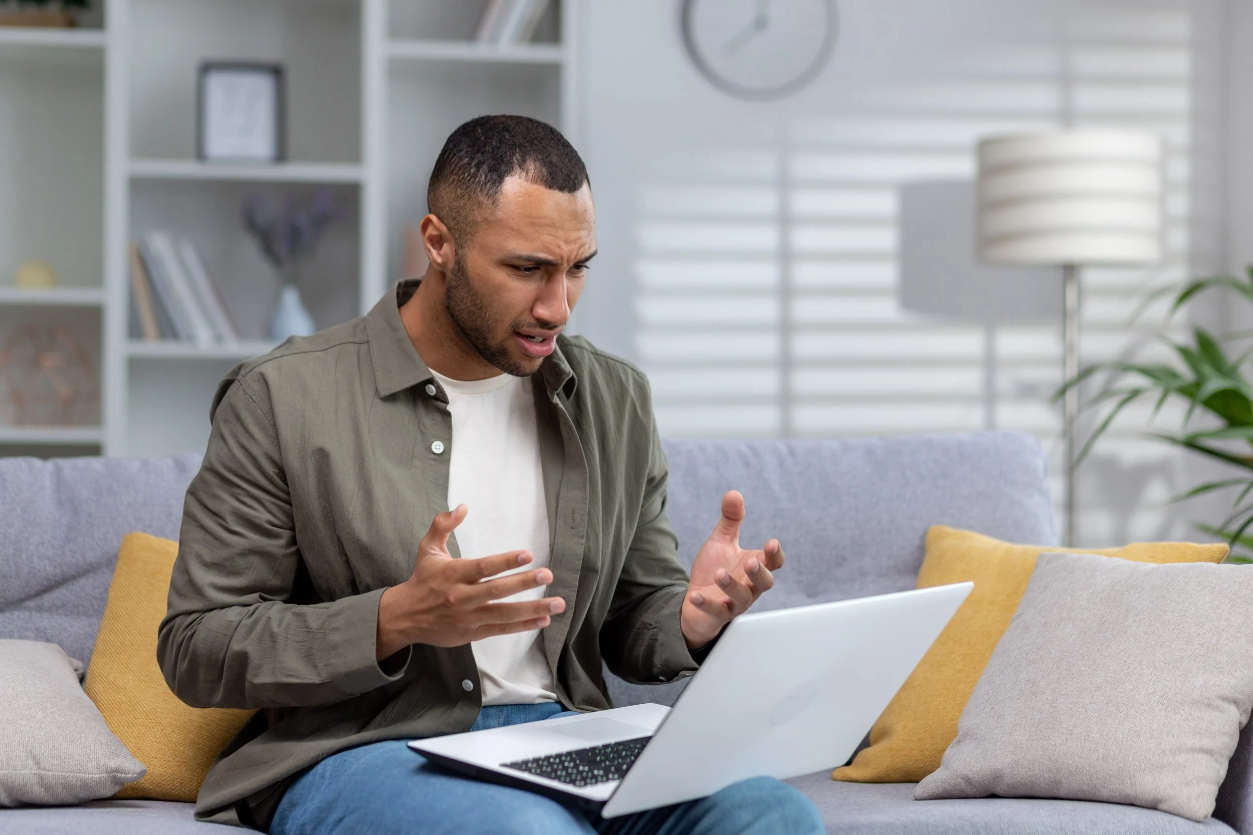 A man sitting on a couch looking confused or frustrated while using a laptop.
