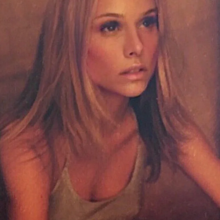 A natural looking attractive woman with long blonde hair, wearing a beige tank top, gazing thoughtfully to the side with a neutral expression.