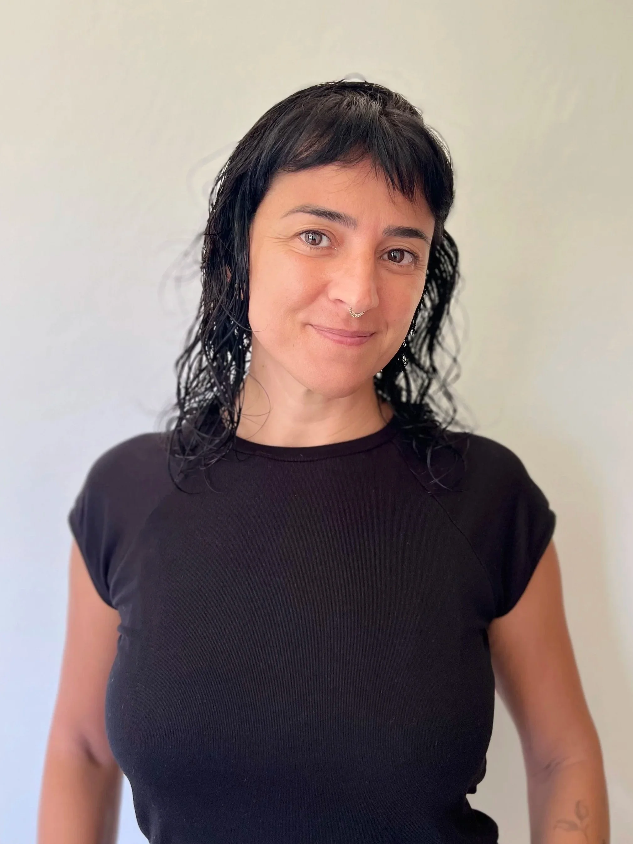 Mujer con cabello negro rizado, camiseta negra, sonrisa suave, fondo claro.