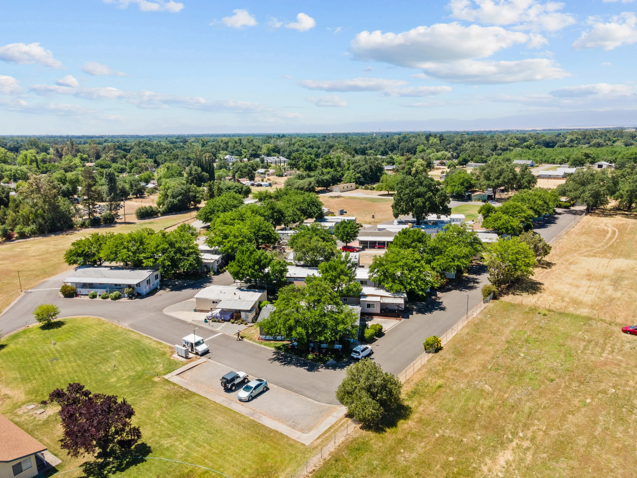  Aerial view of New Orchard Mobile Home & RV Park in Los Molinos, CA, featuring tree-lined streets, parked cars, and open RV spaces with full hookups surrounded by scenic open fields under a partly cloudy Northern California sky — showcasing peaceful