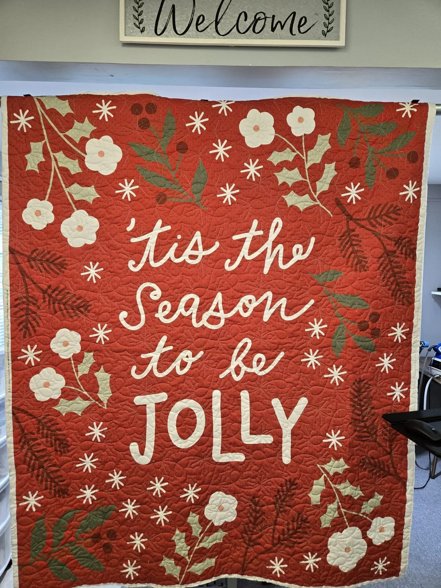 A holiday-themed quilt with a red background decorated with white flowers, green leaves, and holly, featuring the phrase "’tis the season to be JOLLY" in white script.