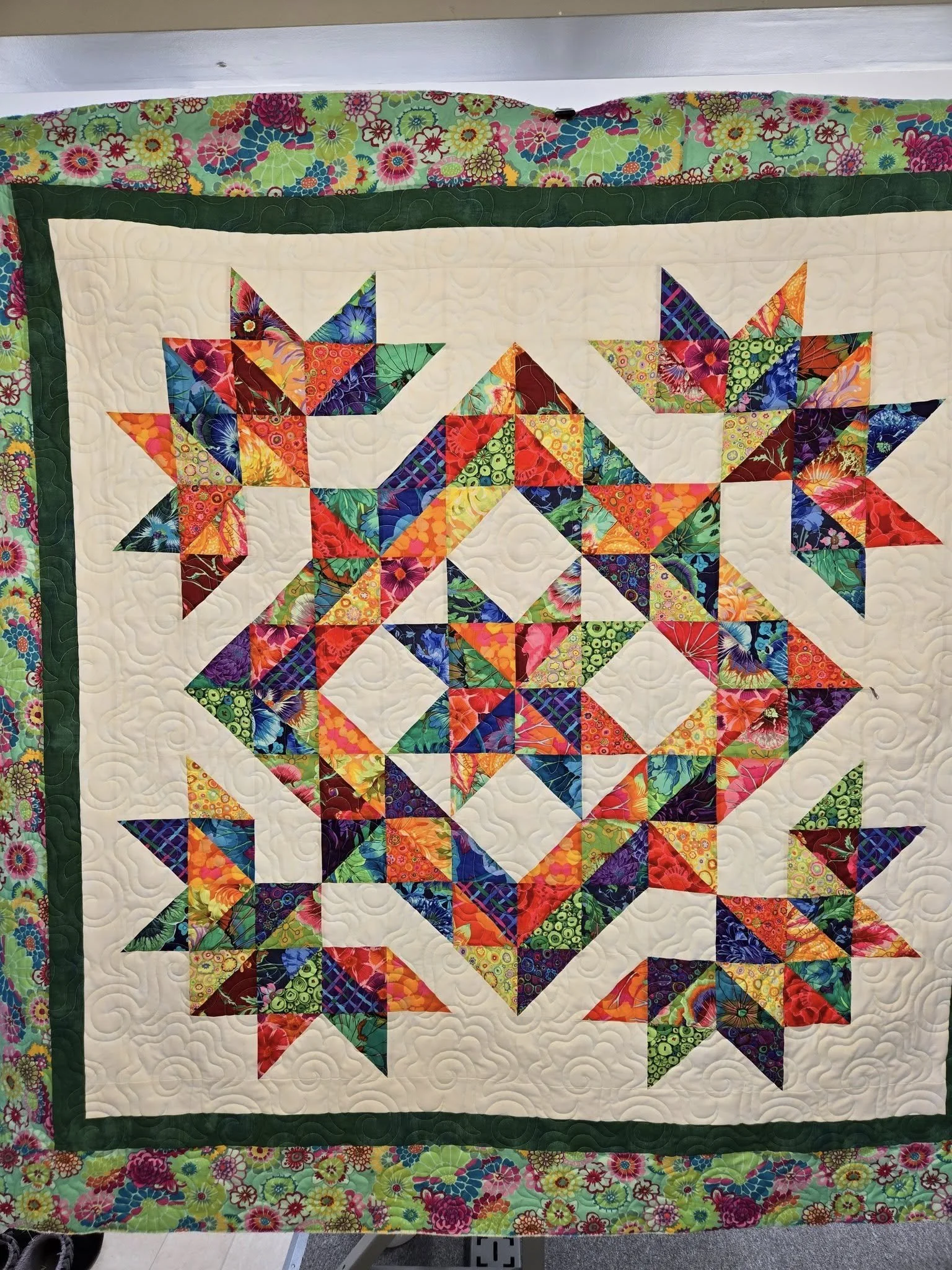A colorful quilt with a central star pattern surrounded by smaller stars, featuring bright, floral, and geometric fabric patches, framed with a floral border.