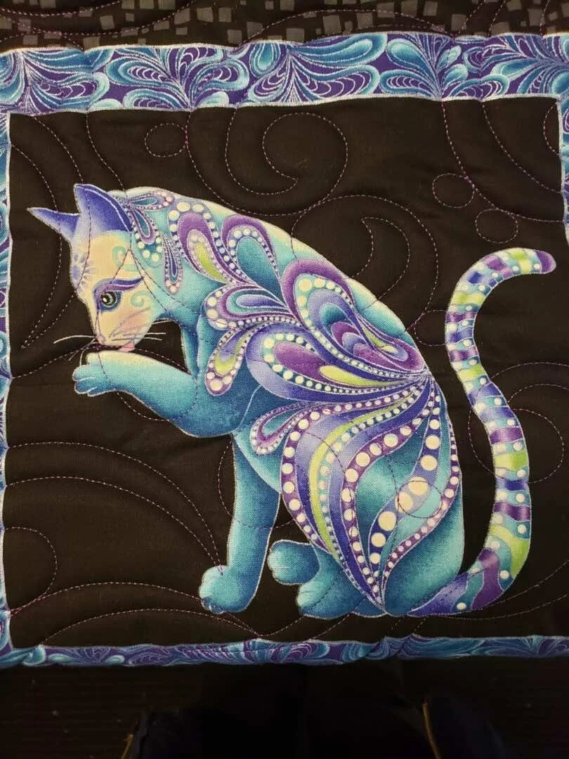 Colorful embroidered artwork of a sitting cat with butterfly wings, on a black quilted fabric background with decorative stitching.