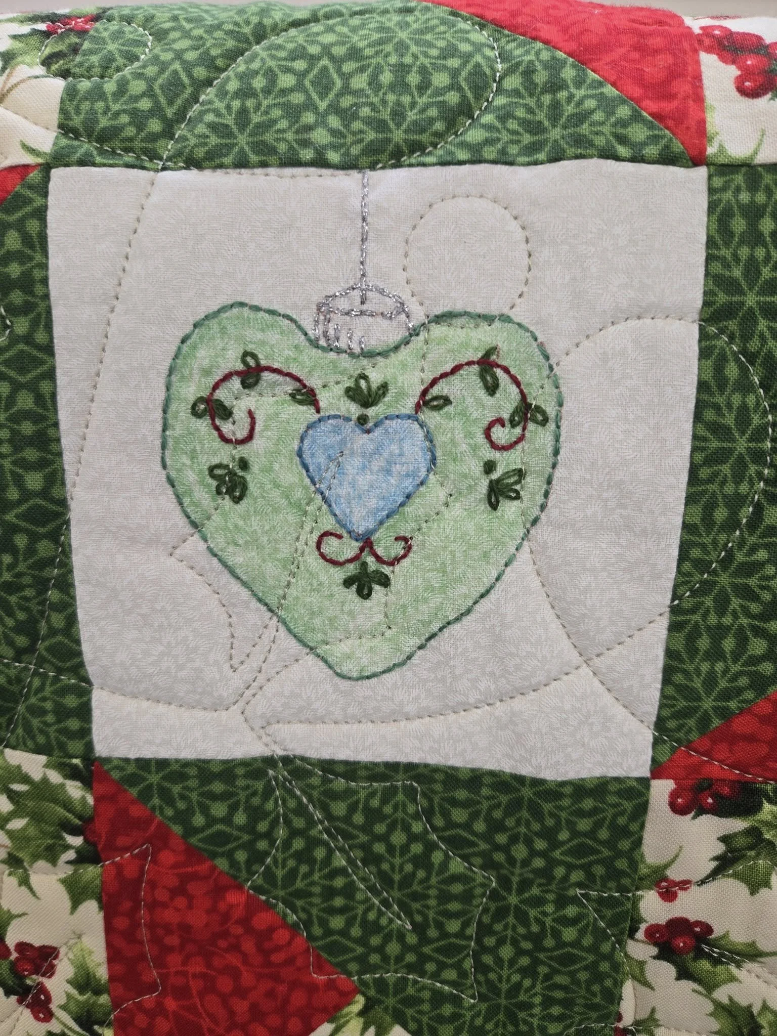 Close-up of a quilted Christmas-themed fabric with an embroidered ornament in the center. The ornament is heart-shaped with a blue heart inside a green heart, decorated with red and green embroidery. Surrounding fabric includes holly leaves and berri