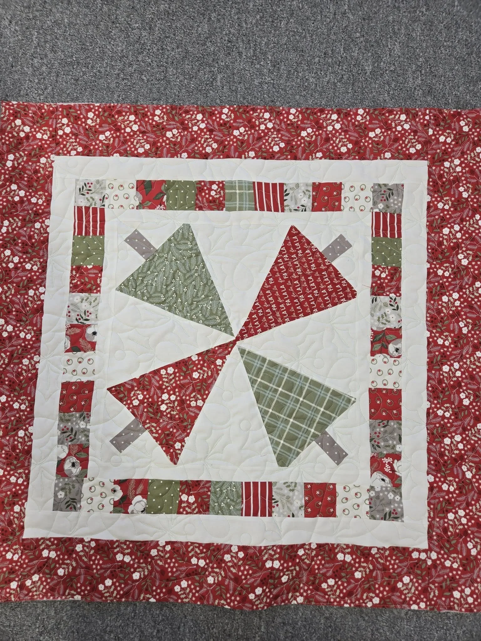 A Christmas-themed quilt with red, green, gray, and white fabric, featuring star patterns and a pieced border, placed on a gray carpet.