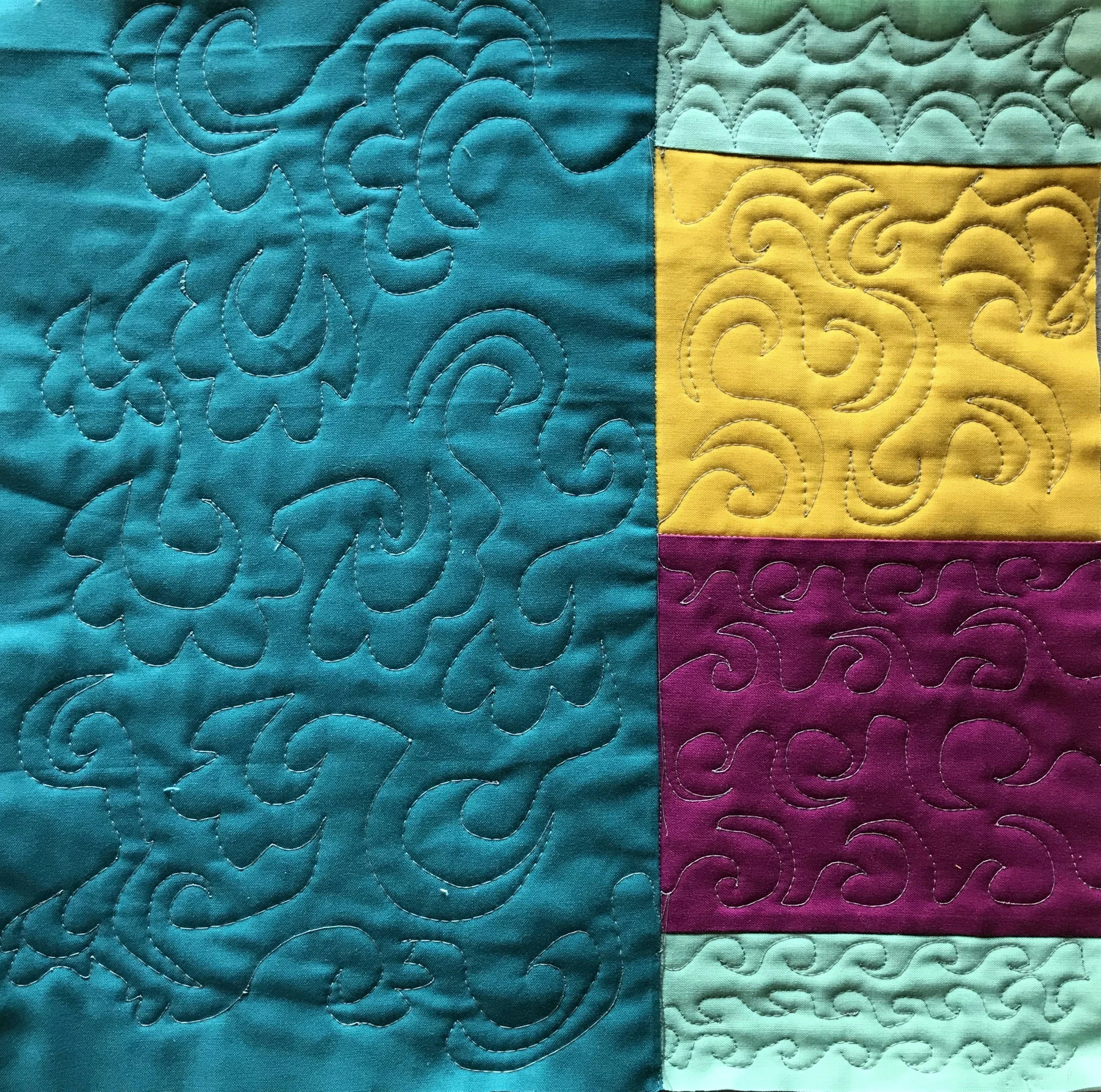 Close-up of a colorful quilt with intricate quilting patterns. The quilt has sections in teal, yellow, purple, and light green, each featuring swirling stitch designs.