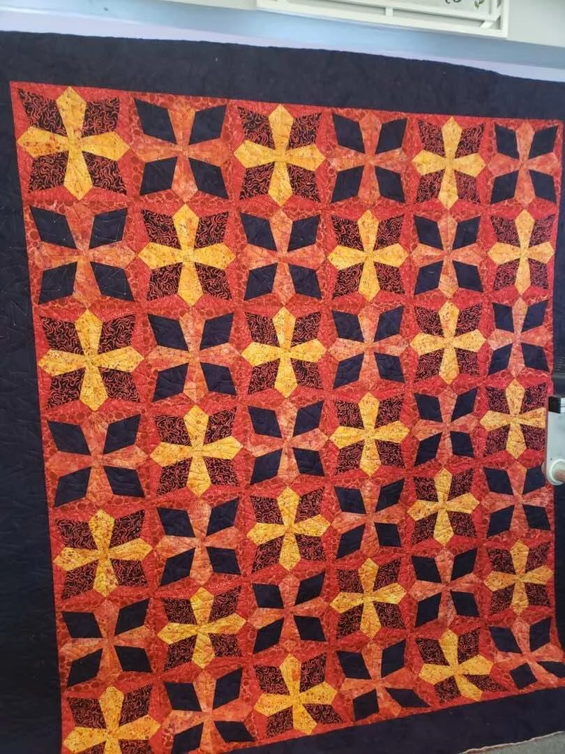 A quilt with a geometric pattern in red, yellow, and black fabric, displayed on a wall.