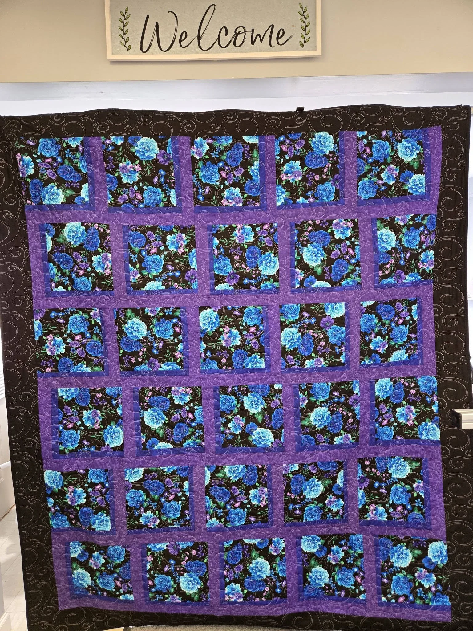 A quilt with a floral pattern of blue and purple flowers on a black background, bordered by purple fabric, hanging on a wall with a sign above that reads "Welcome" with decorative leaves.