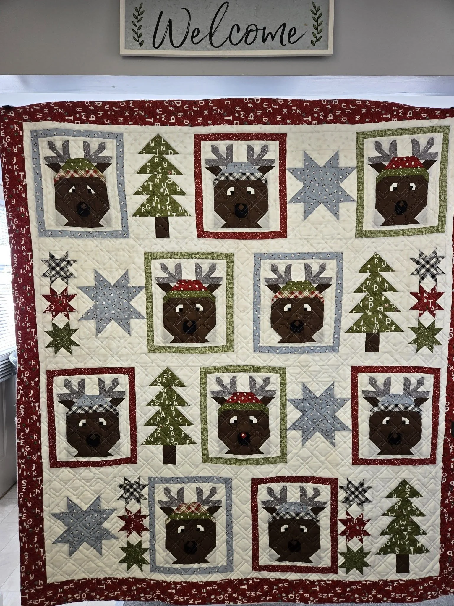 A quilt with Christmas themed designs, including reindeer with red and green accessories, snowflakes, and Christmas trees, hanging on a wall with a "Welcome" sign above it.