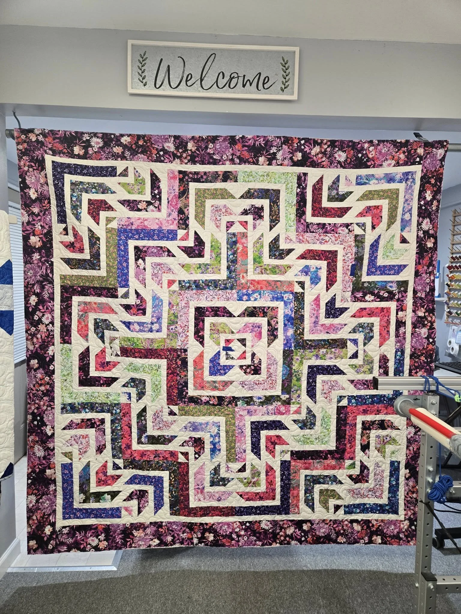 A colorful floral quilt with a geometric spiral pattern hanging on a wall, with a 'Welcome' sign above it.
