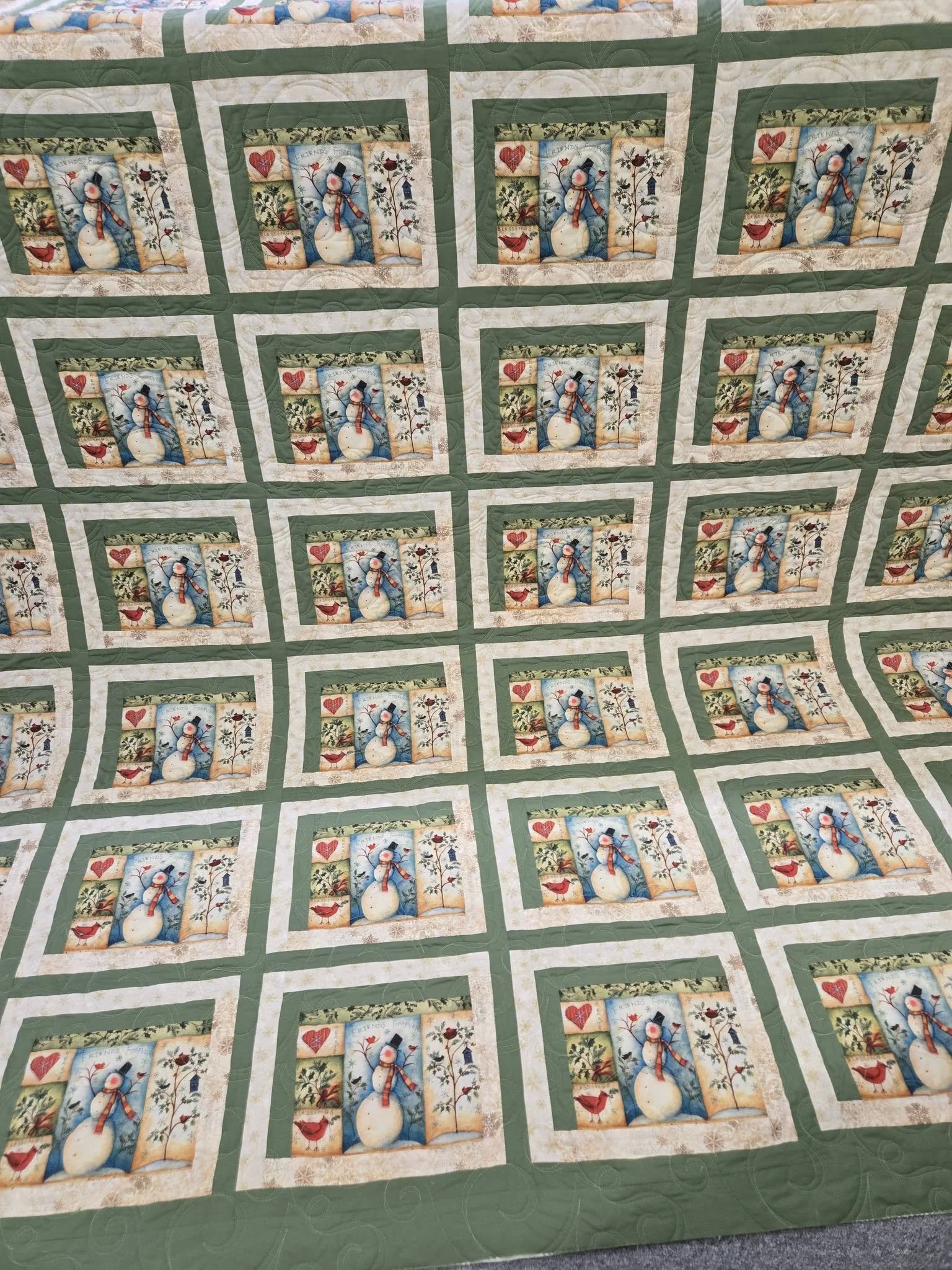 A quilt with a pattern of snowmen, hearts, trees, and birds in a winter scene, bordered by green and white fabric.