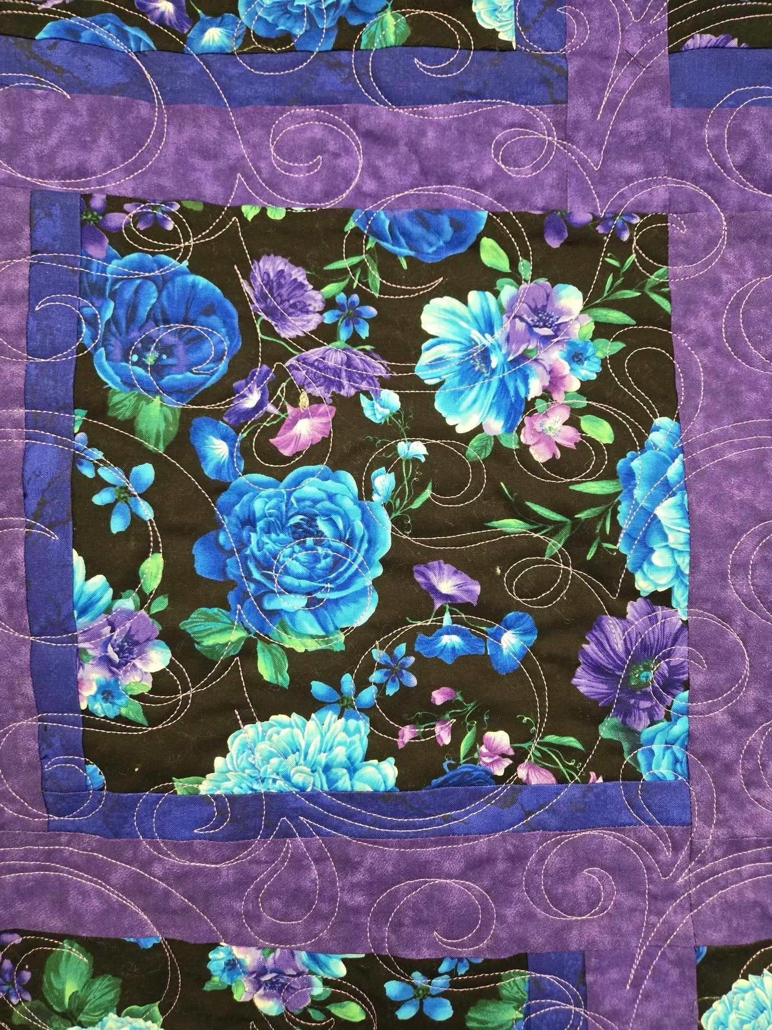 Close-up of a quilt with a black background floral pattern featuring blue, purple, and pink flowers, bordered by purple fabric with decorative stitching.