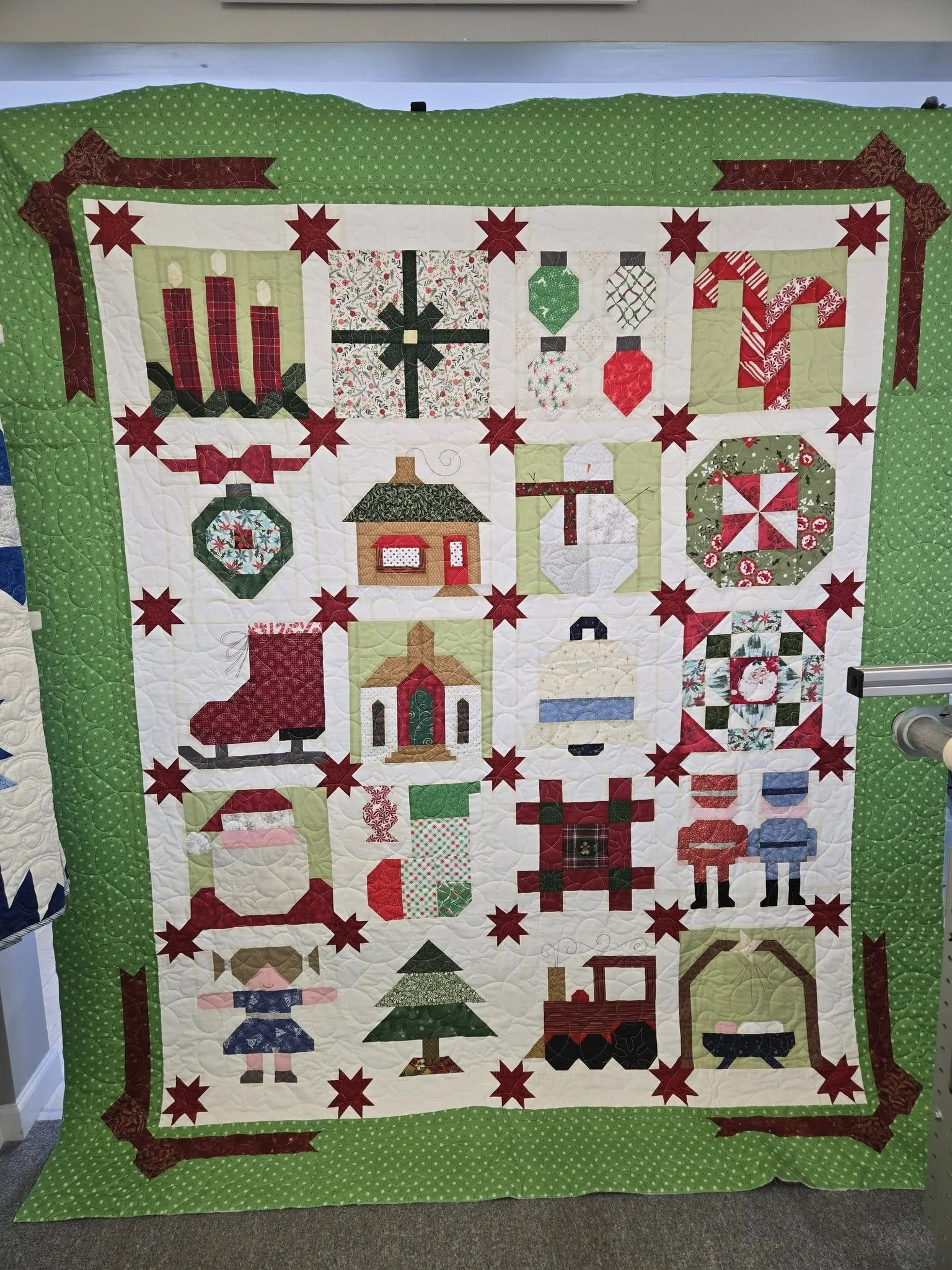 A holiday-themed quilt with various Christmas motifs, including candles, ornaments, a house, a Christmas tree, a Santa figure, and a train, all bordered by a green polka dot pattern.