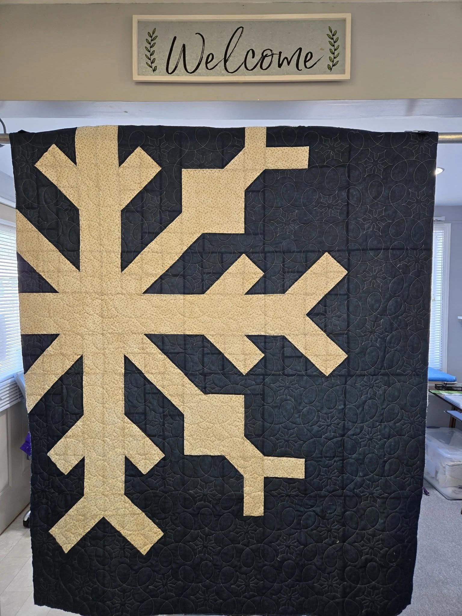 A quilt with a large beige snowflake pattern on a black background, hanging in front of a wall with a framed 'Welcome' sign on top.