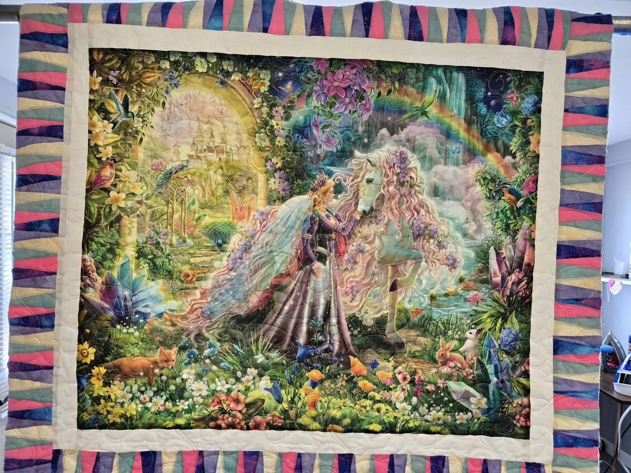 A colorful quilt with a fantasy scene depicting a woman with long flowing hair riding a white unicorn in a lush garden with animals, flowers, rainbow, and a distant castle.