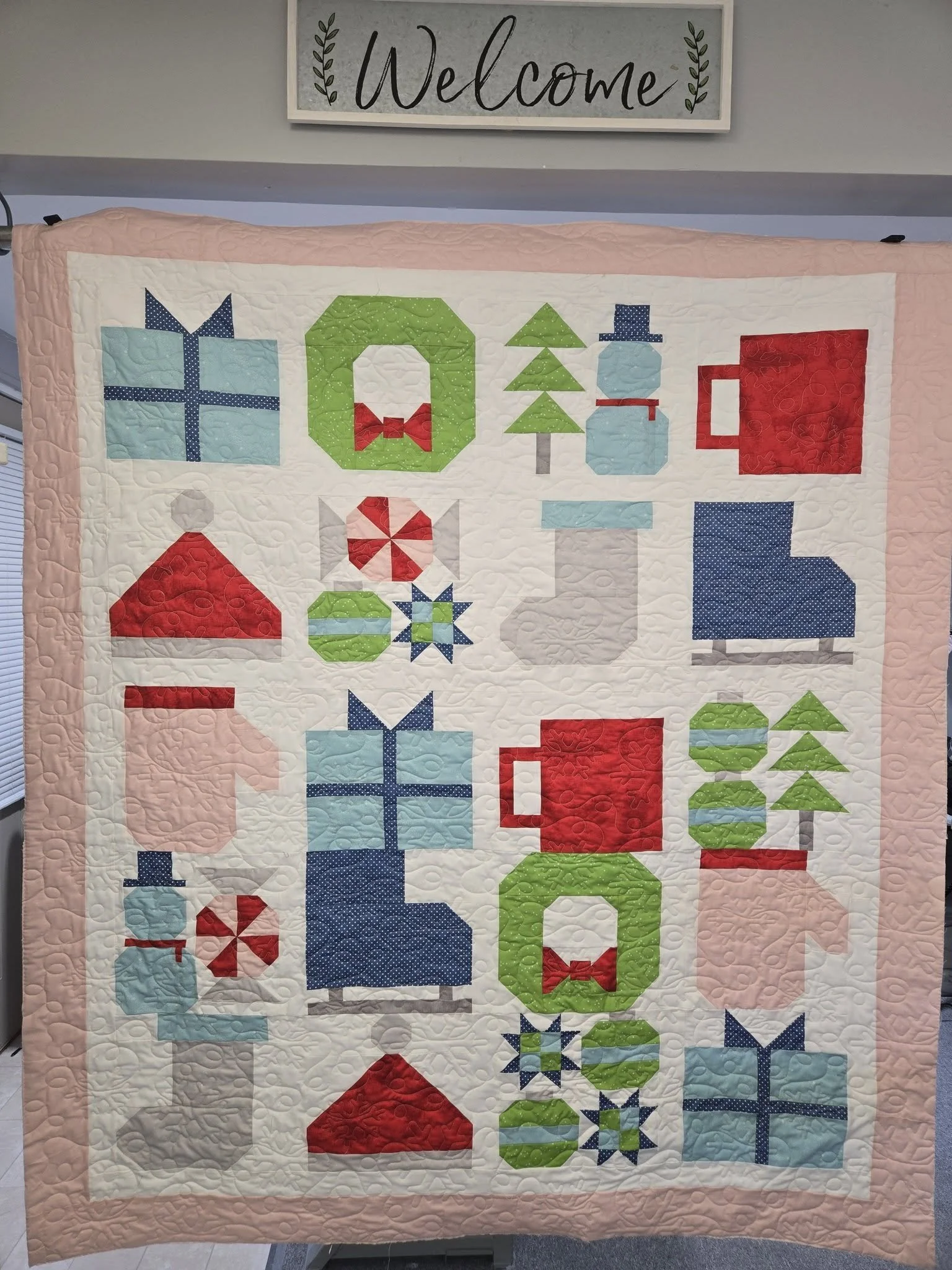 A quilt with Christmas-themed blocks including gifts, a tree, a snowman, a Santa hat, a stocking, and ornaments, on a pink border. Above the quilt, there is a sign that says "Welcome" with leafy decorations.