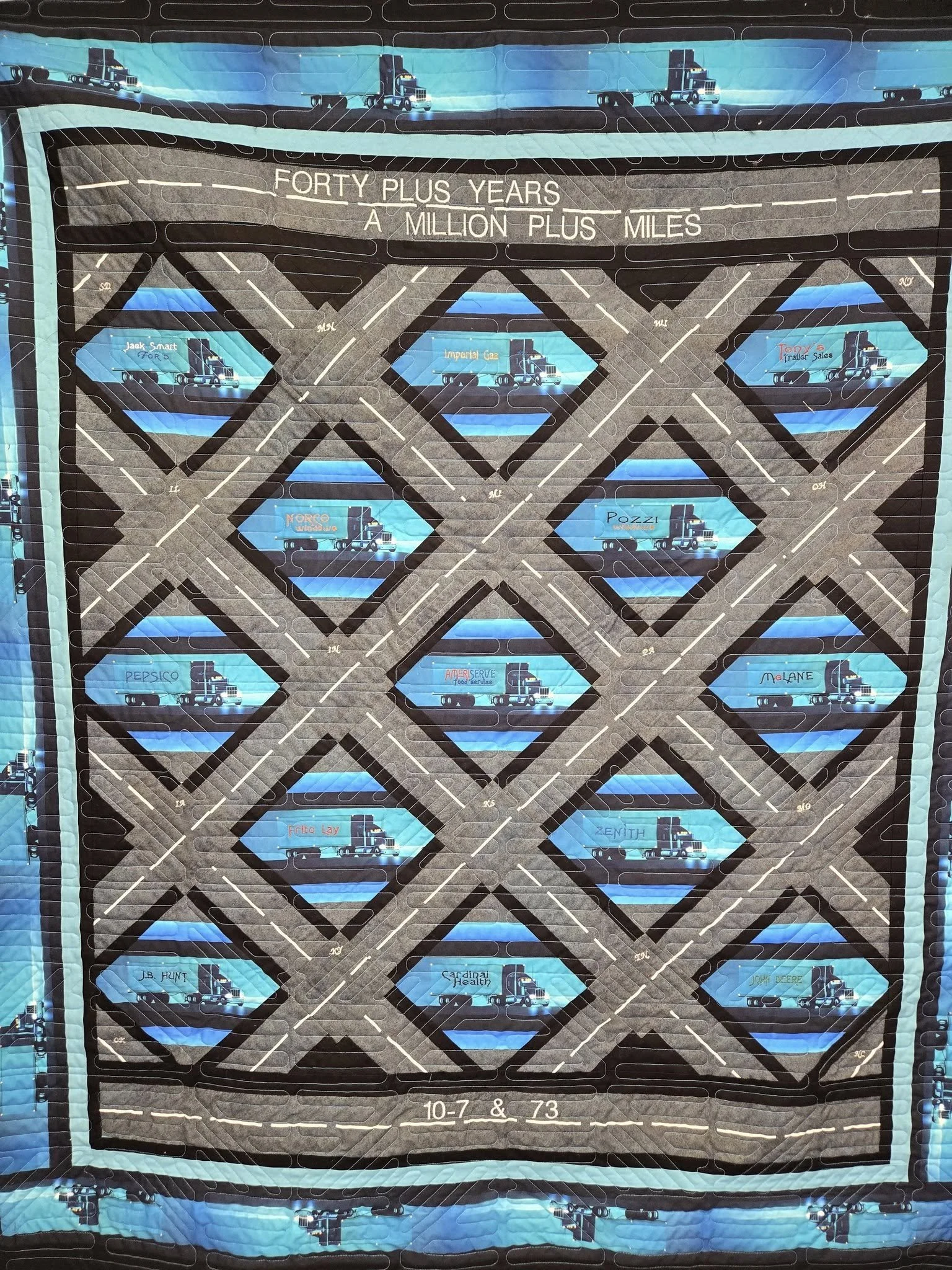 A quilt with a highway theme featuring trucks and road signs, with the text 'Forty Plus Years, A Million Plus Miles' and a date '10-7 & 73' at the bottom.