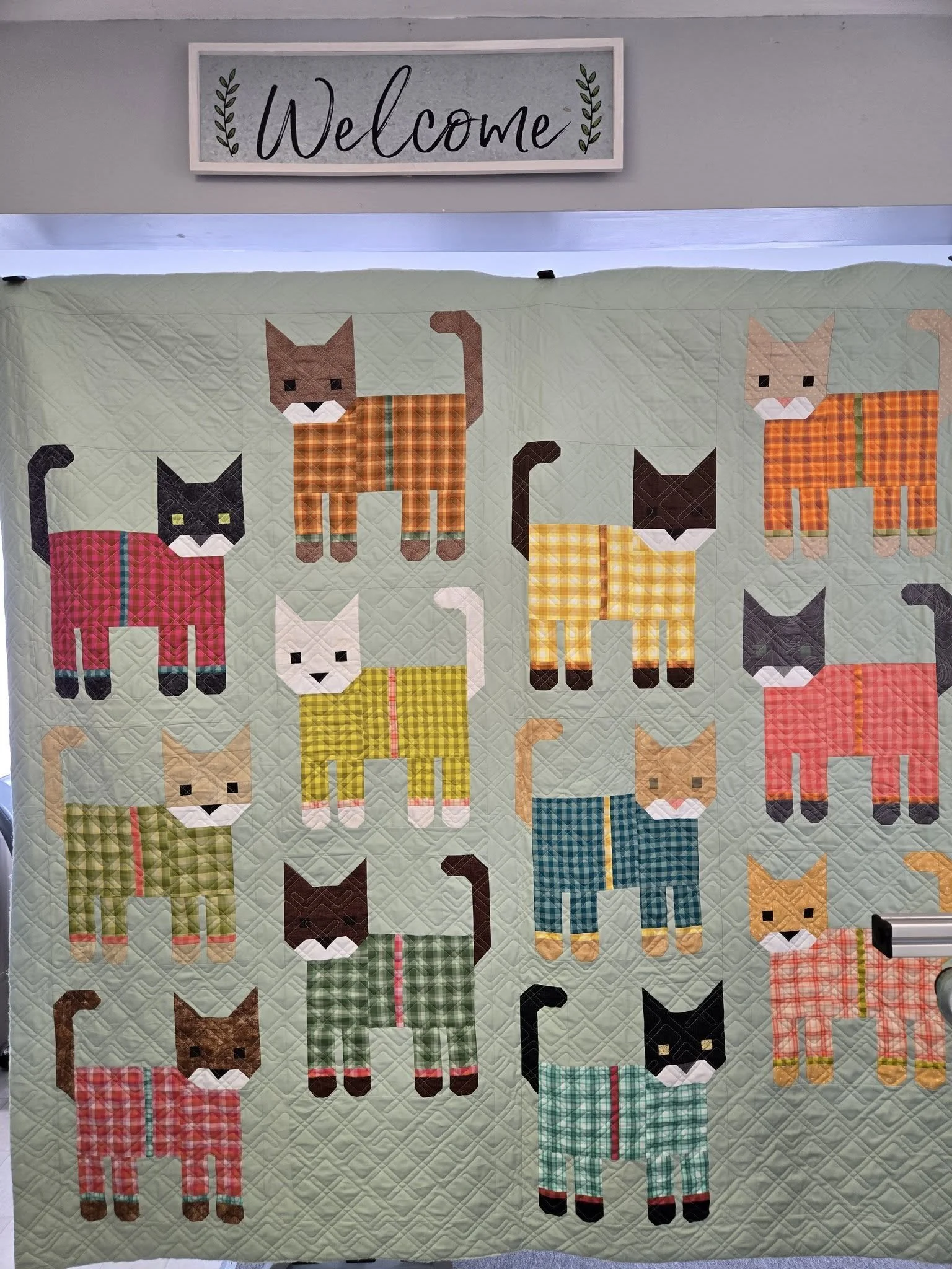 A quilt with colorful plaid cats on a pale green background and a 'Welcome' sign above it.