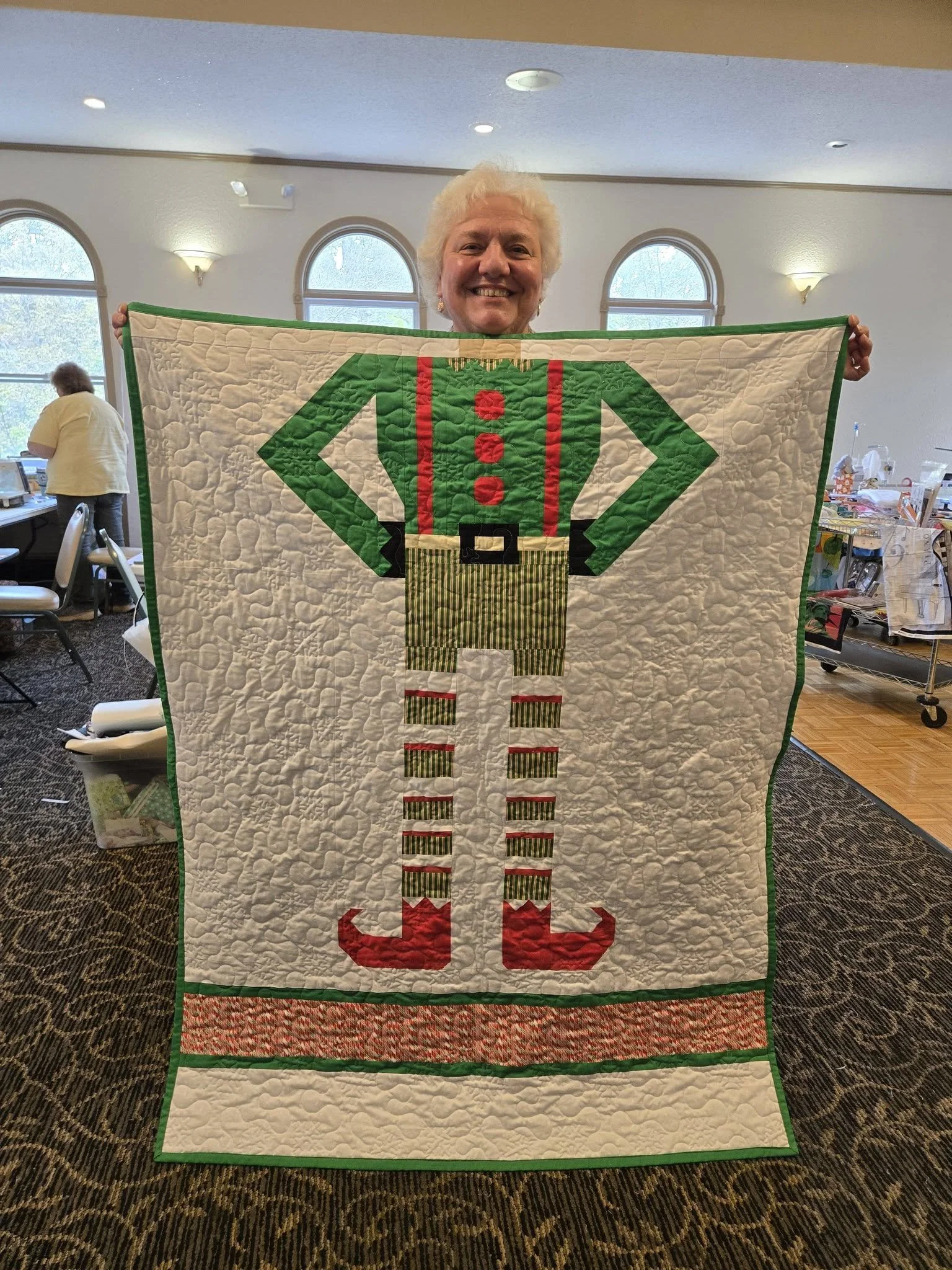 A woman with white hair smiling while holding up a Christmas-themed quilt featuring an elf design in a room with large arched windows.