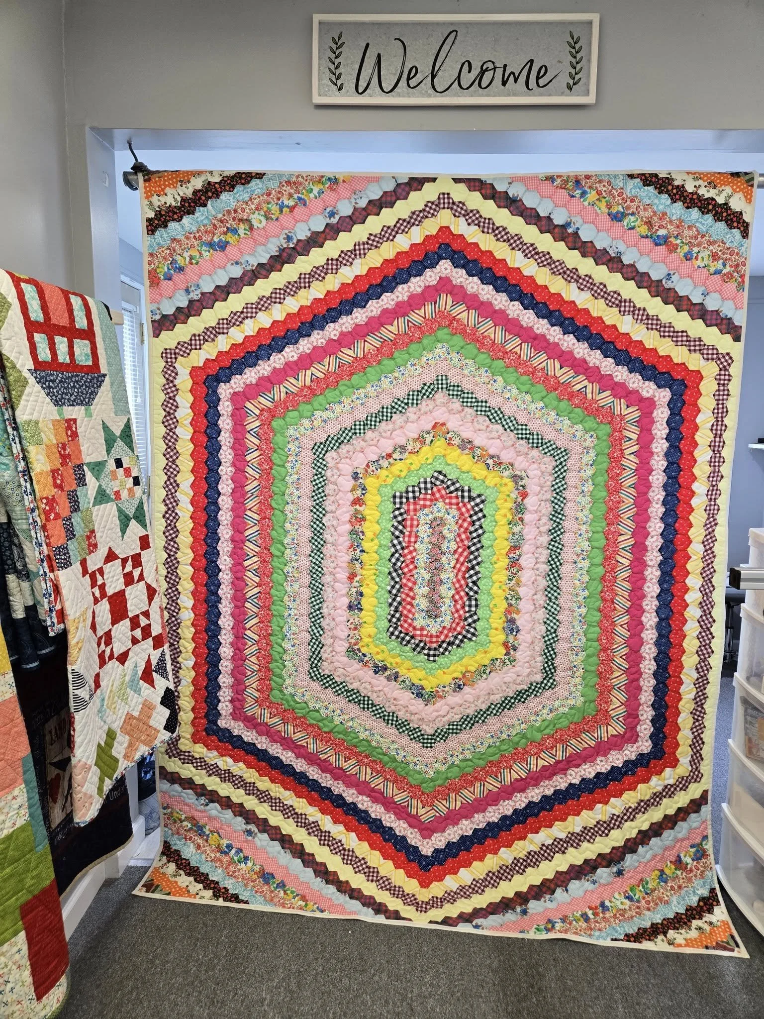 Colorful patchwork quilt with a hexagon pattern hanging on a wall with a welcome sign above it in a craft room.