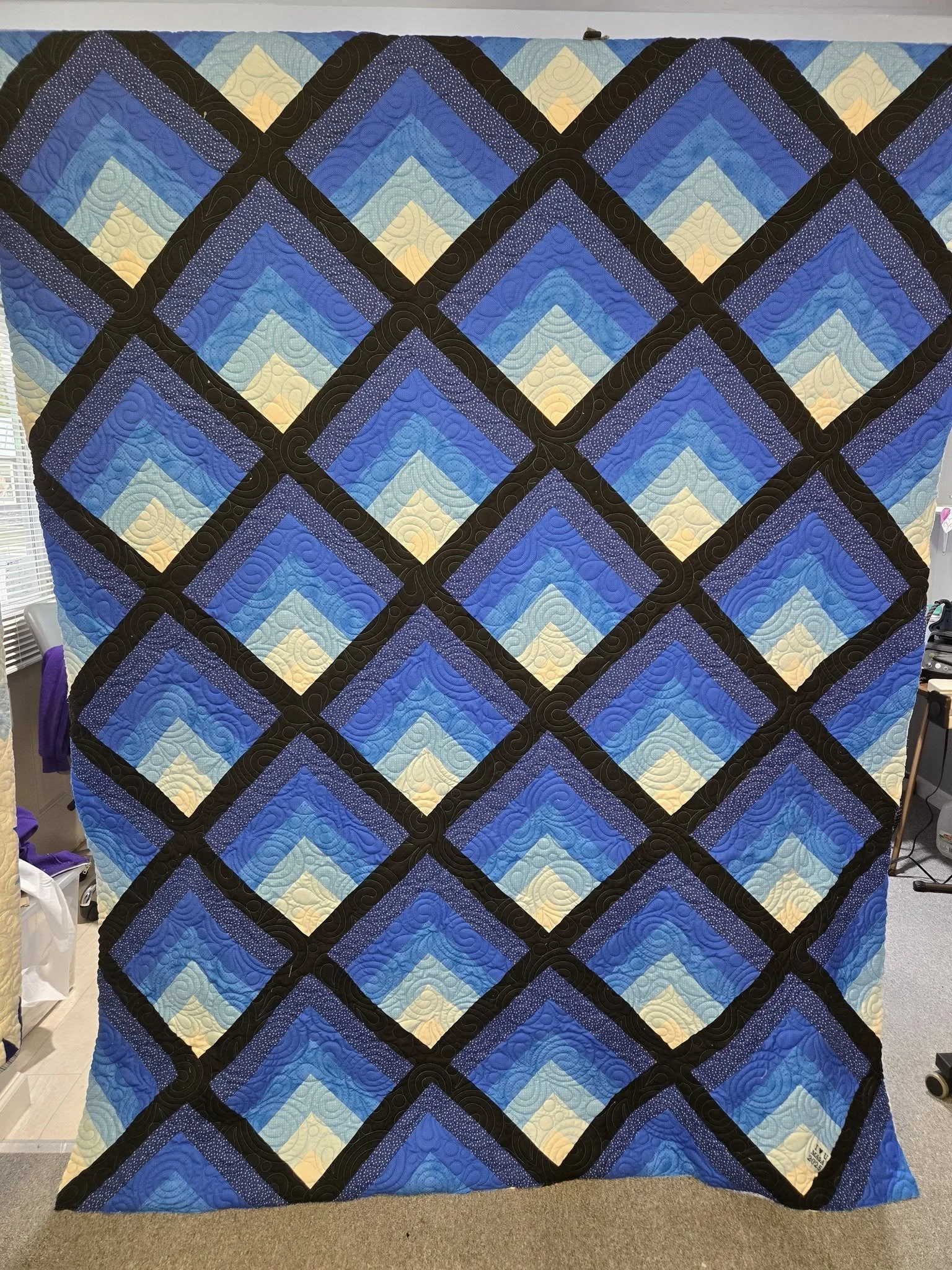 A colorful quilt with a geometric diamond pattern in blue, yellow, purple, and black fabric. The quilt is displayed hanging vertically.
