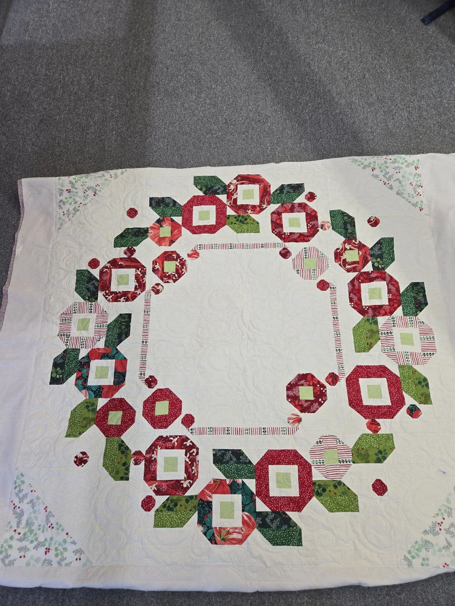 A quilt with a holiday wreath pattern made of fabric hexagons, squares, and leaf shapes in red, green, and patterned fabrics, arranged on a white background.