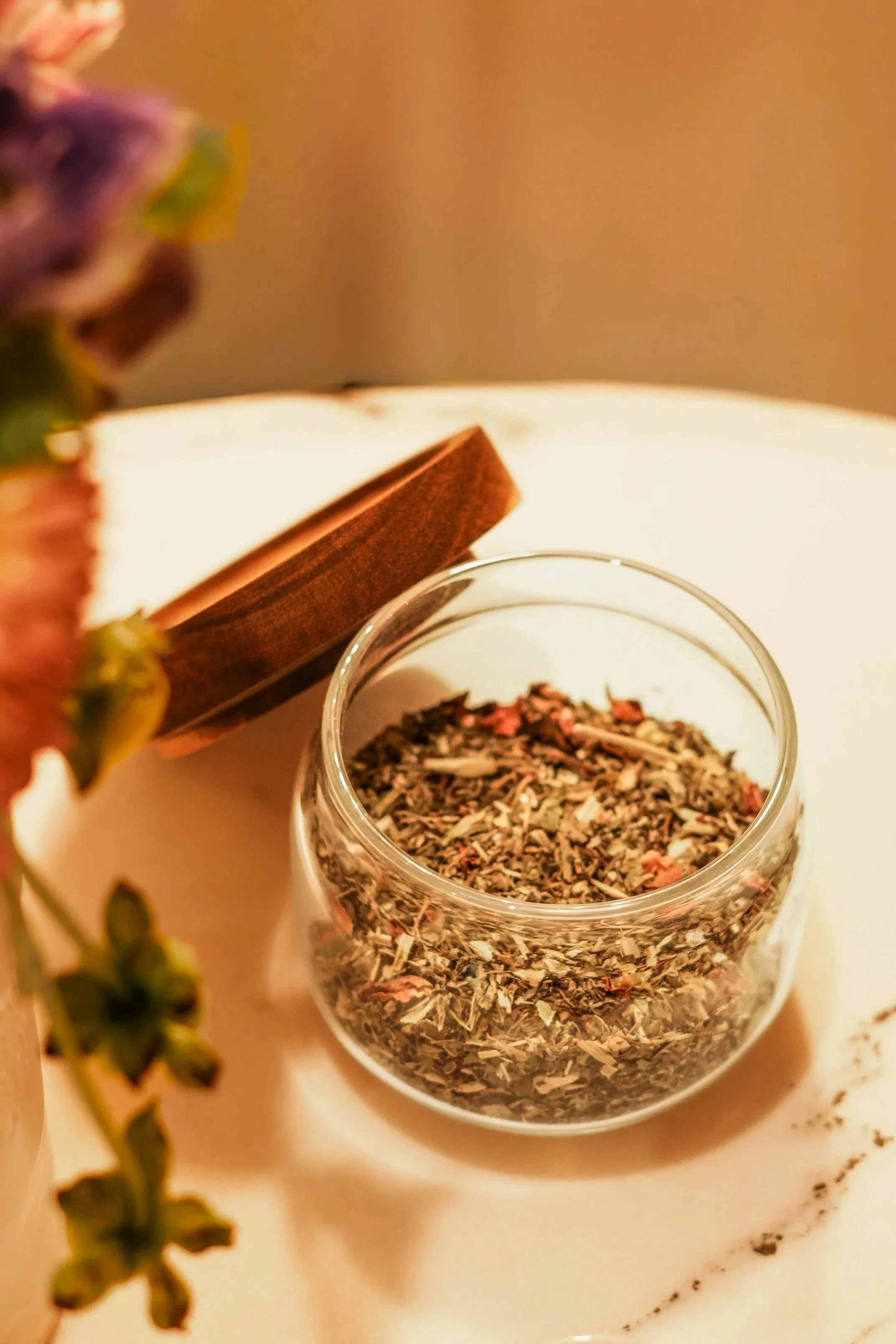 Close-up of a glass jar filled with dried herbal tea leaves on a white surface, with an open wooden box and blurred purple flowers in the background. Raspberry Leaf Tea blend.
